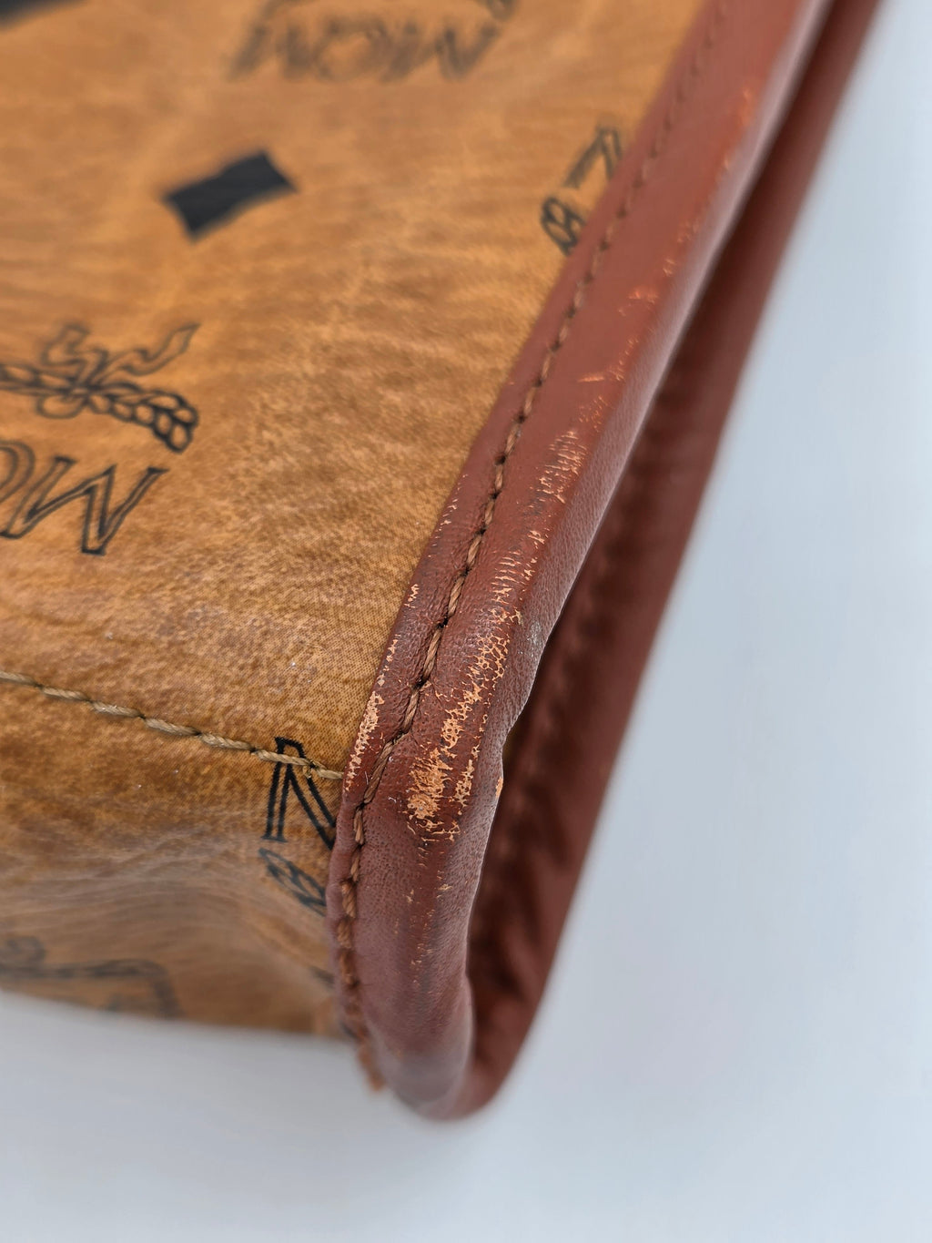 Close-up of a vintage MCM canvas pouch showing the tan coated canvas body with black logo print and a distressed brown leather trim along the edge.