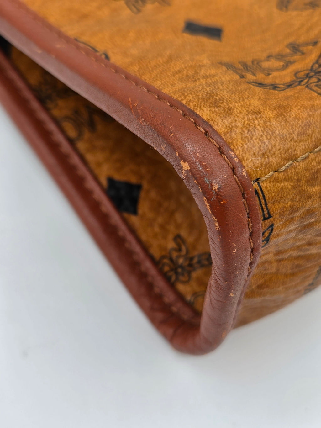 Close-up of a vintage MCM monogram canvas pouch or wallet corner, showing the tan-brown coated canvas with black printed logo motifs and a contrasting brown leather trim on a white background.
