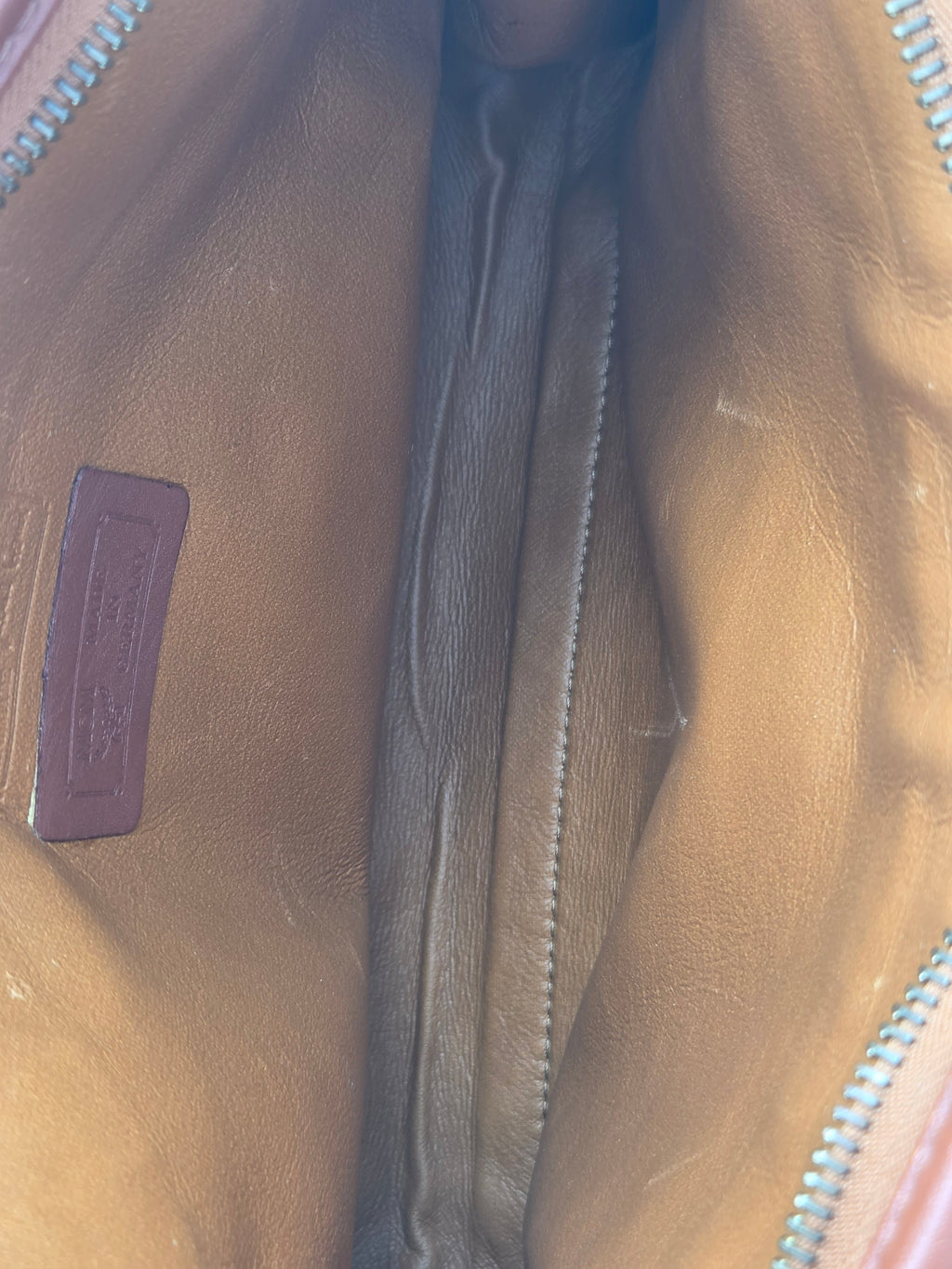 Close-up of the interior lining of a tan leather bag, showing a zippered opening, stitched interior channel, and a small embossed brand patch near the bottom.