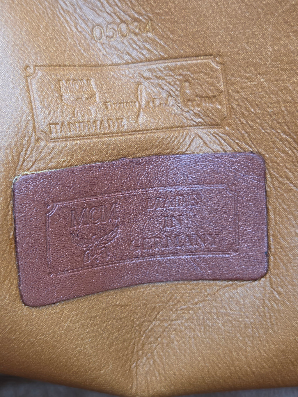 A close-up of a tan leather surface with an embossed rectangular patch and a smaller sewn brown leather brand patch reading 'Vogue' and 'Gentleman' with a floral emblem.