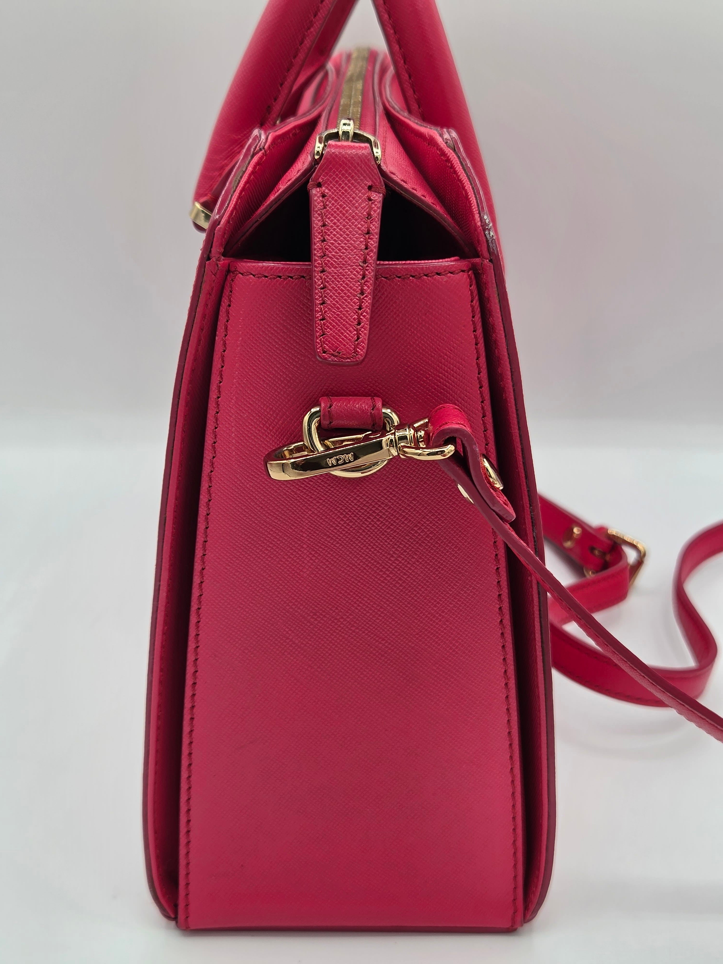 A close-up of a structured handbag in a rich magenta pink leather with gold-tone hardware and a matching shoulder strap, shown against a plain white background.