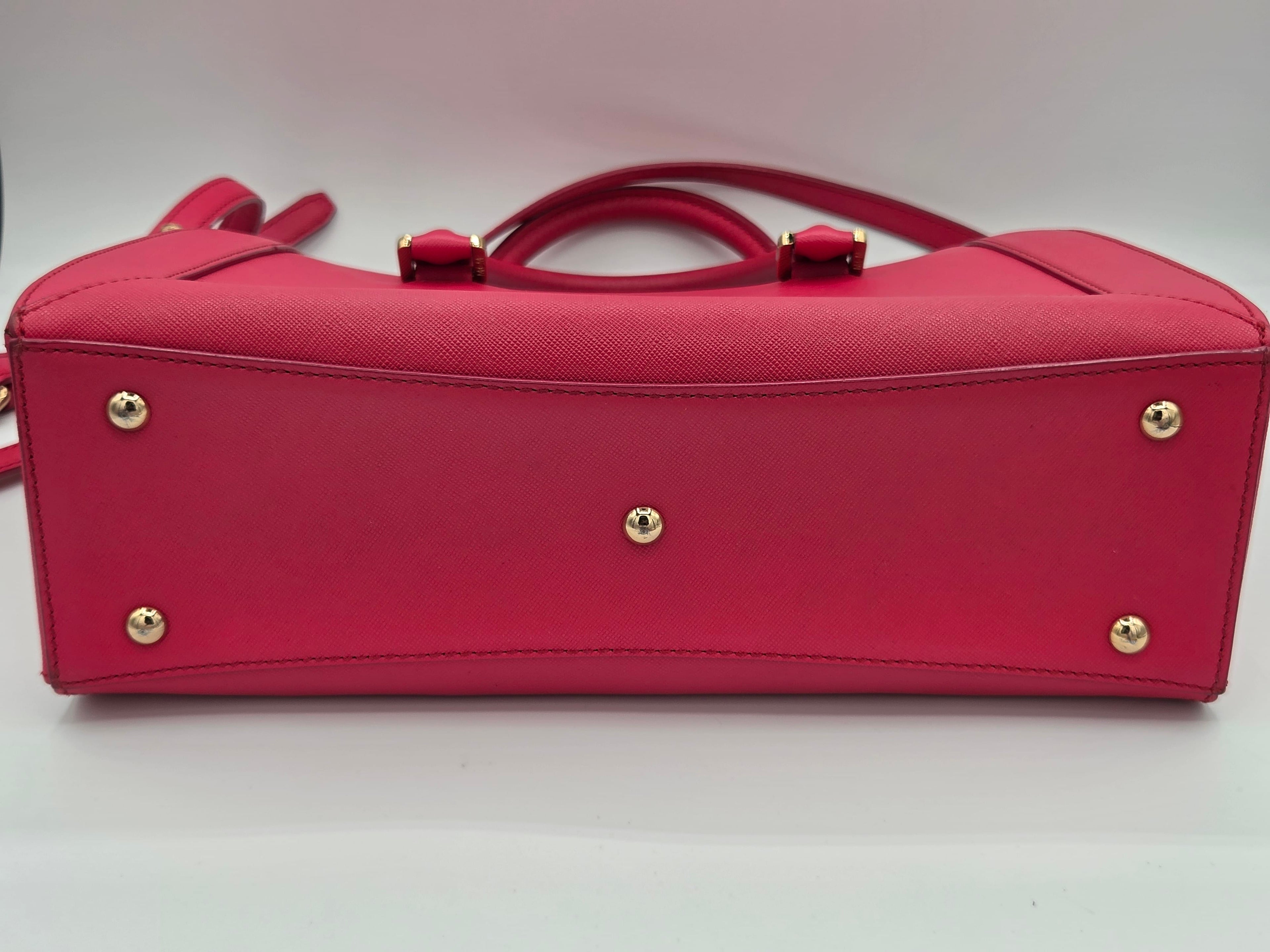 A bright pink textured leather handbag shown from the bottom, featuring gold-tone metal studs and a top handle, on a white background.