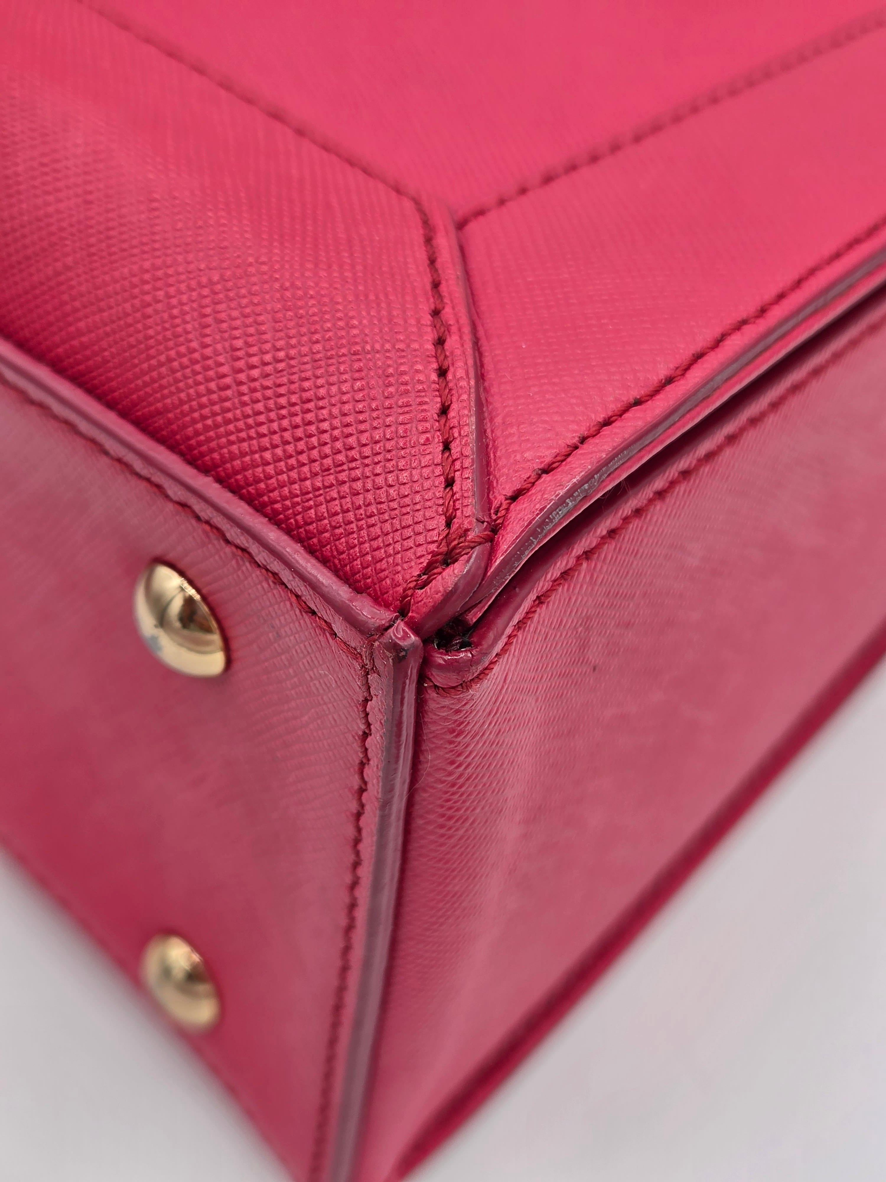 Close-up detail of a pink textured leather handbag corner, showing precise stitching, a brass grommet, and a gold-tone metal rivet on a light gray background.