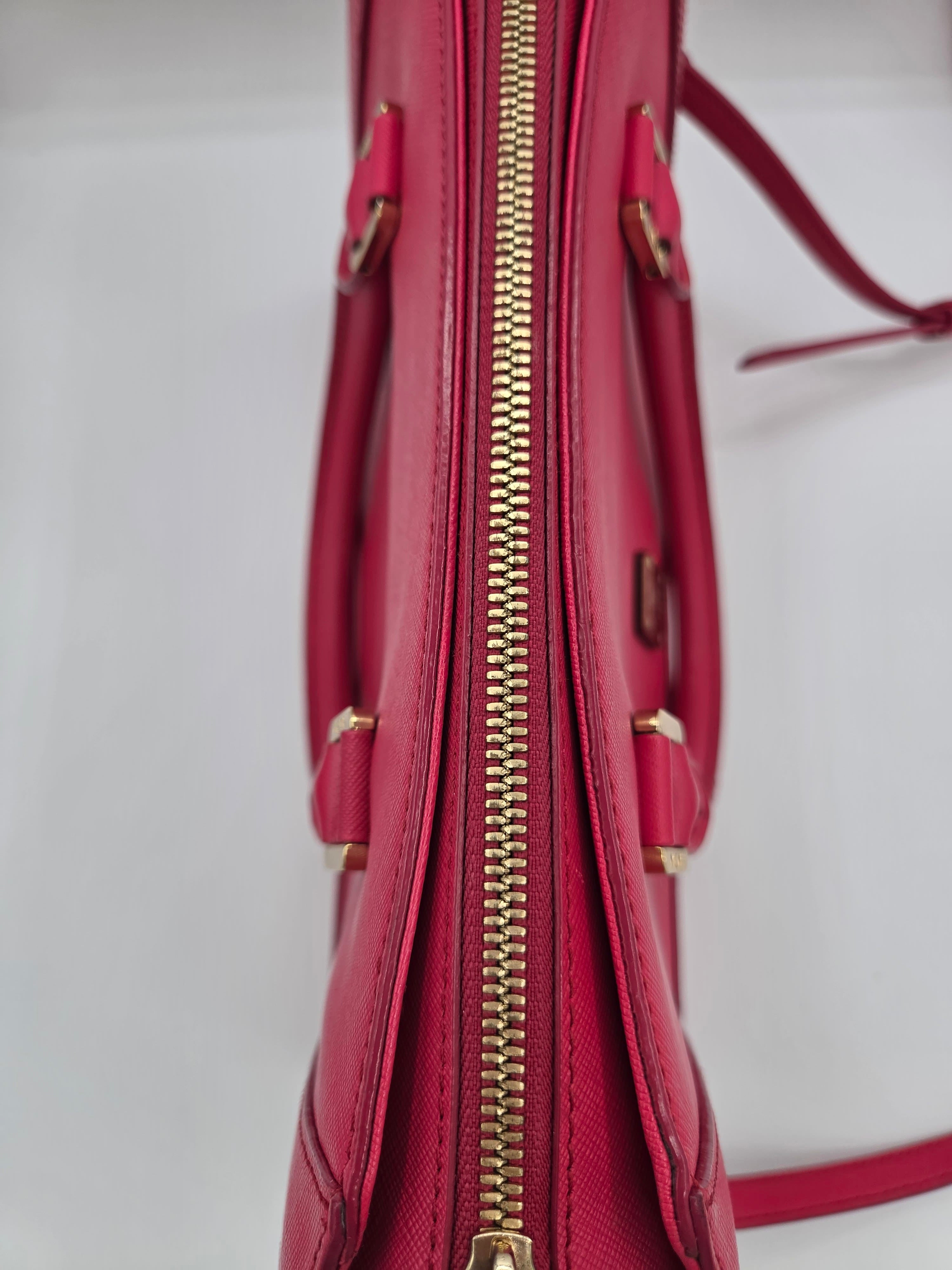 A close-up of a red leather handbag showing the side profile with a gold zipper, top handles, and a matching red shoulder strap against a plain white background.