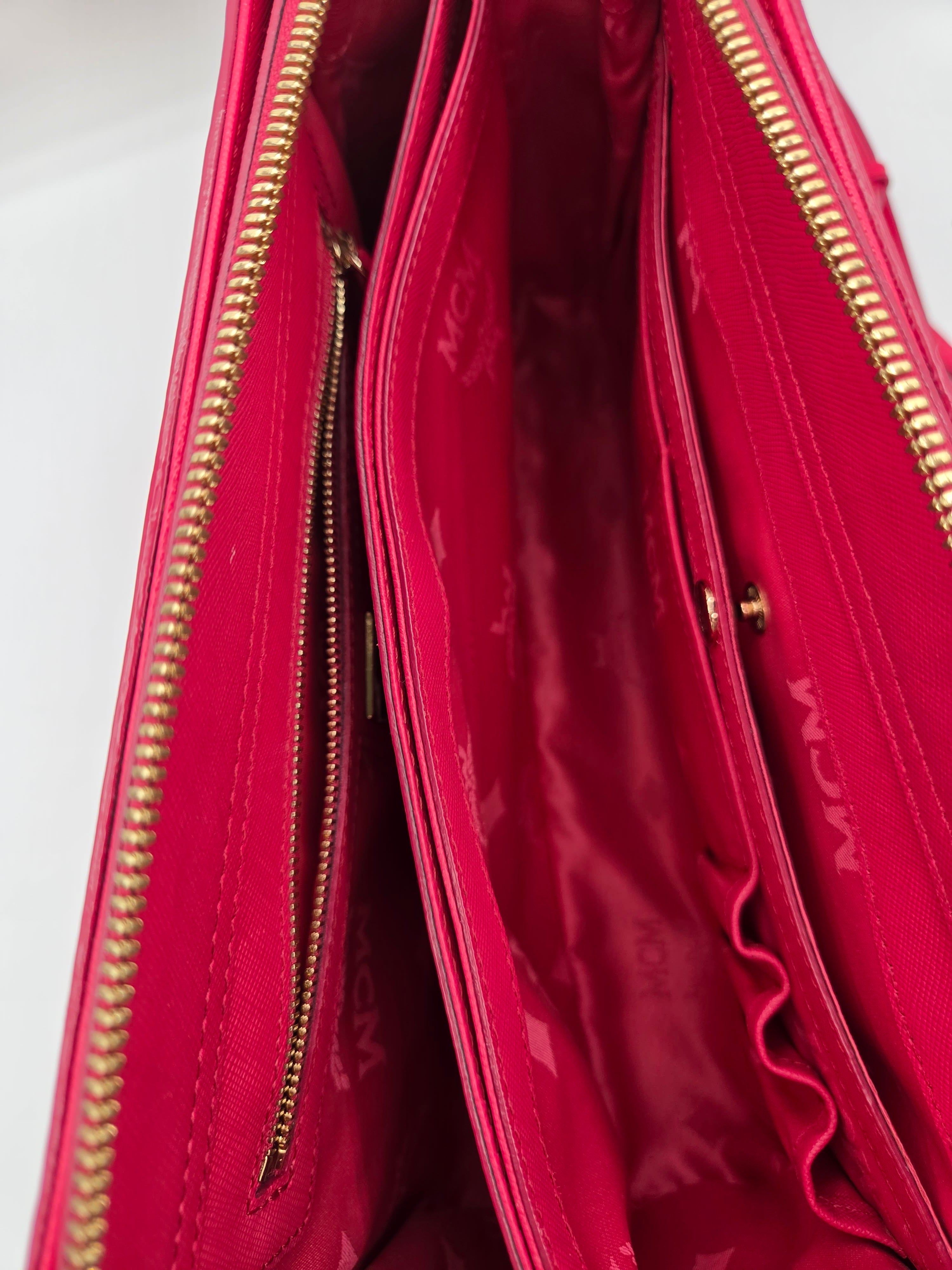 Close-up of the interior of a pink MCM clutch or wallet, showing multiple compartments, gold-tone zippers, and the signature MCM logo pattern on the lining.