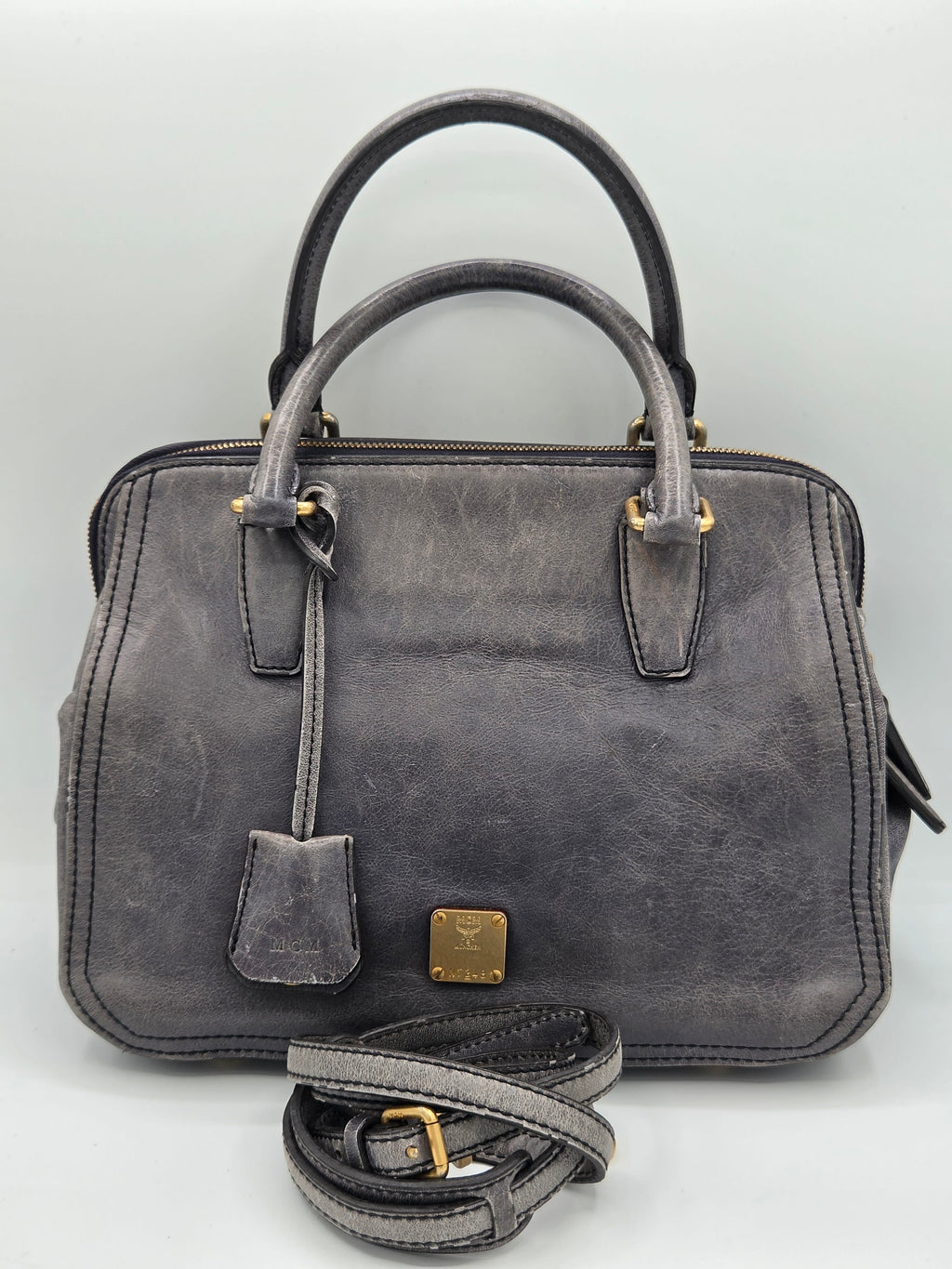 A distressed dark gray leather handbag with gold-tone hardware, featuring dual top handles, a front hanging tag, and a matching detachable shoulder strap coiled in front, set against a plain light gray background.