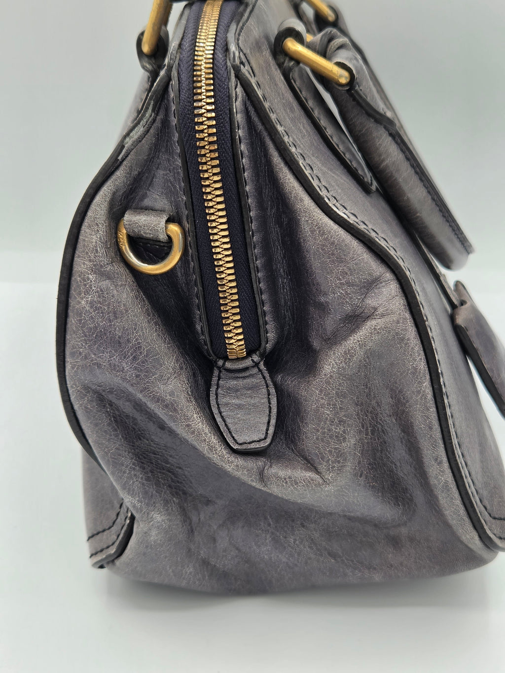 A close-up of a distressed gray leather handbag showing a gold zipper, gold hardware ring, and dark stitching details against a light gray background.