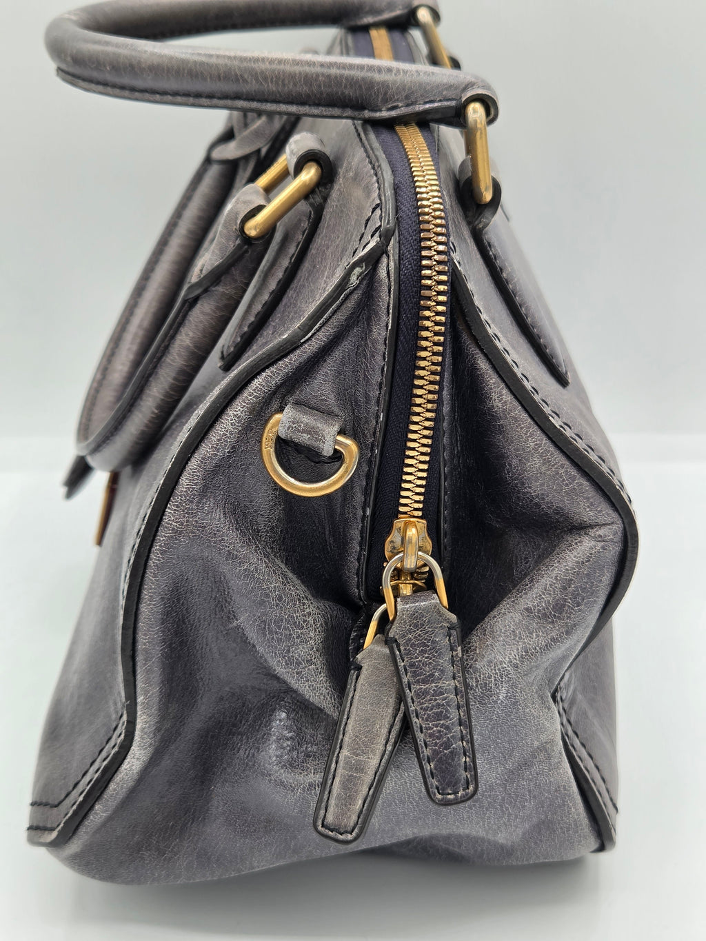 A close-up of a dark gray leather handbag showing the interior side with a gold zipper, gold-tone hardware, and stitched details.