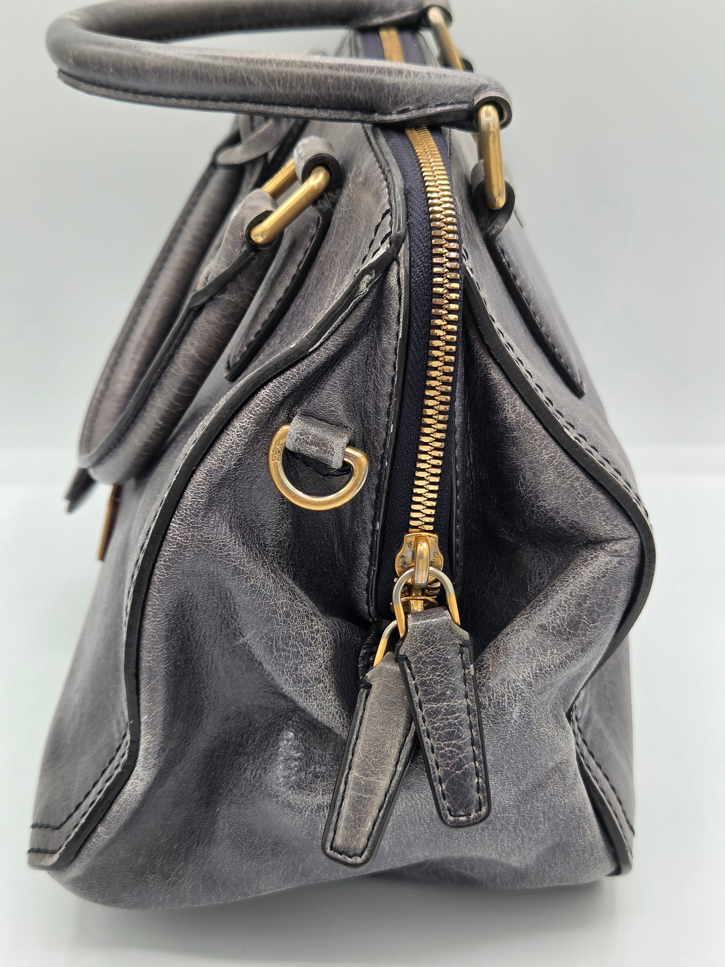 A close-up of a dark gray leather handbag showing the interior side with a gold zipper, gold-tone hardware, and stitched details.