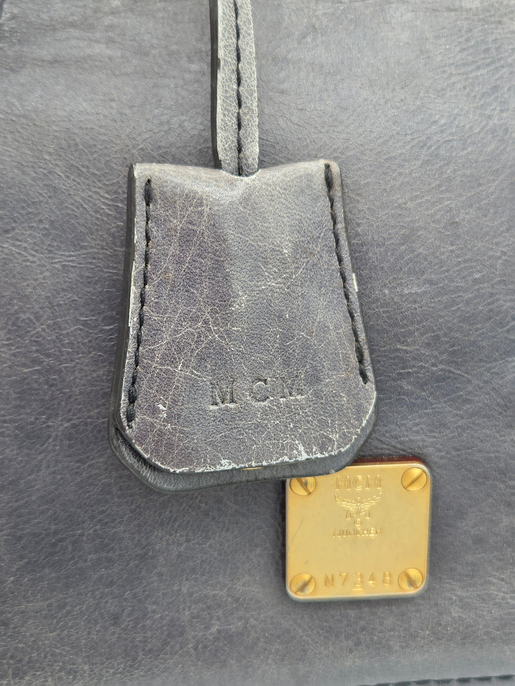 Close-up of a dark gray leather MCM luggage tag attached to a matching gray leather bag, featuring an embossed MCM logo and a gold-tone square metal brand plaque.