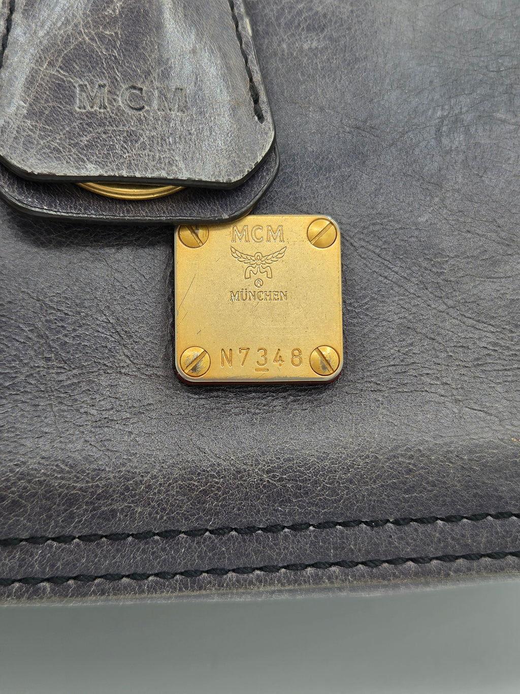 Close-up of a dark gray leather MCM bag featuring a gold-tone square metal plate engraved with 'MCM München' and 'N7348', alongside a matching leather tab with an embossed MCM logo.