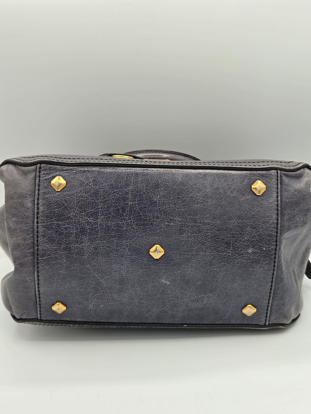 A close-up of a dark gray leather handbag showing the bottom panel with gold-tone metal studs and rivets, set against a neutral gray background.