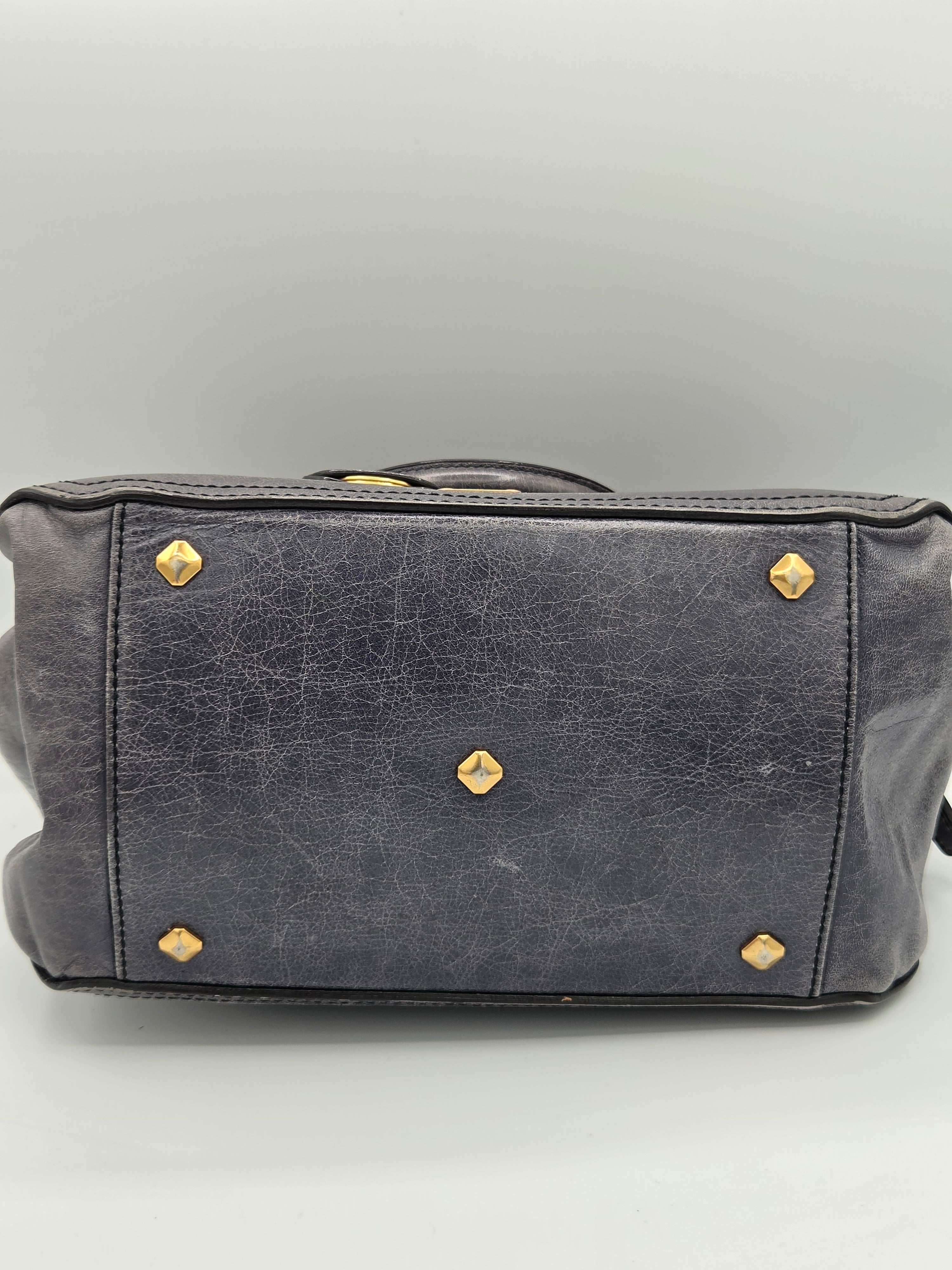 A close-up of a dark gray leather handbag showing the bottom panel with gold-tone metal studs and rivets, set against a neutral gray background.