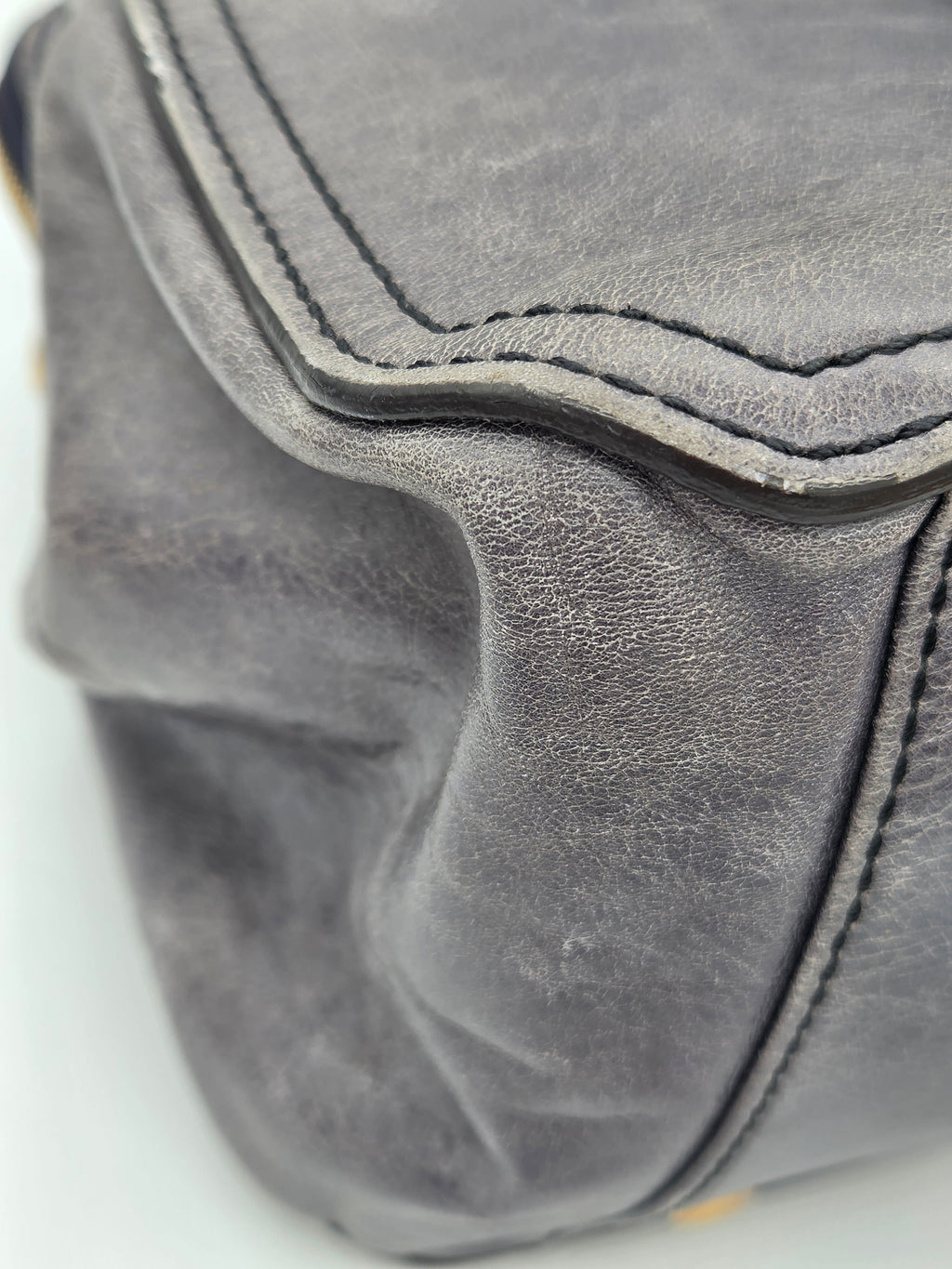 Close-up of a gray leather or faux-leather bag corner showing stitched edges and textured material against a white background.