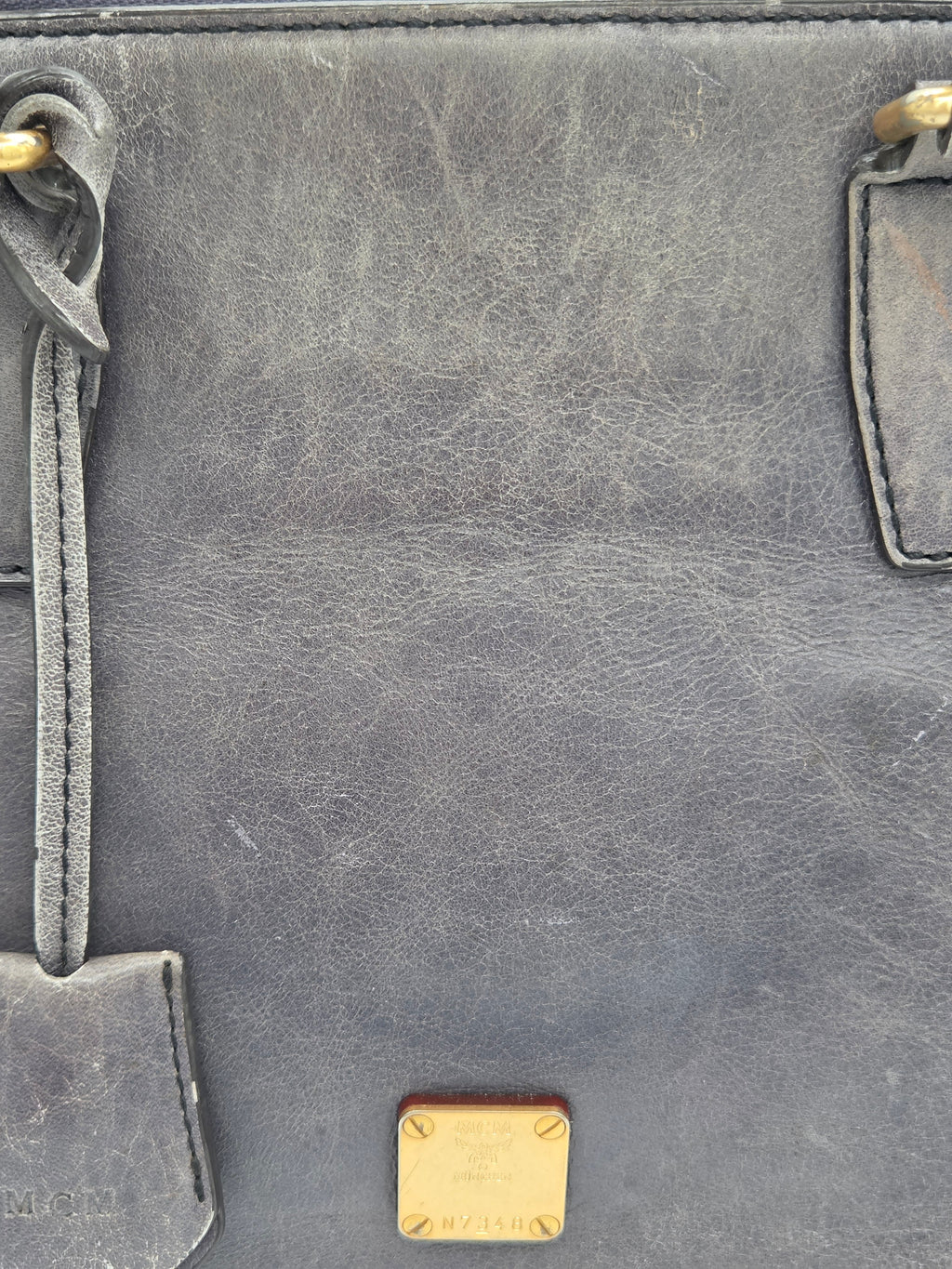 Close-up of a dark gray distressed leather bag showing the textured grain, gold-tone hardware, and a small gold-tone metal logo plaque at the bottom.