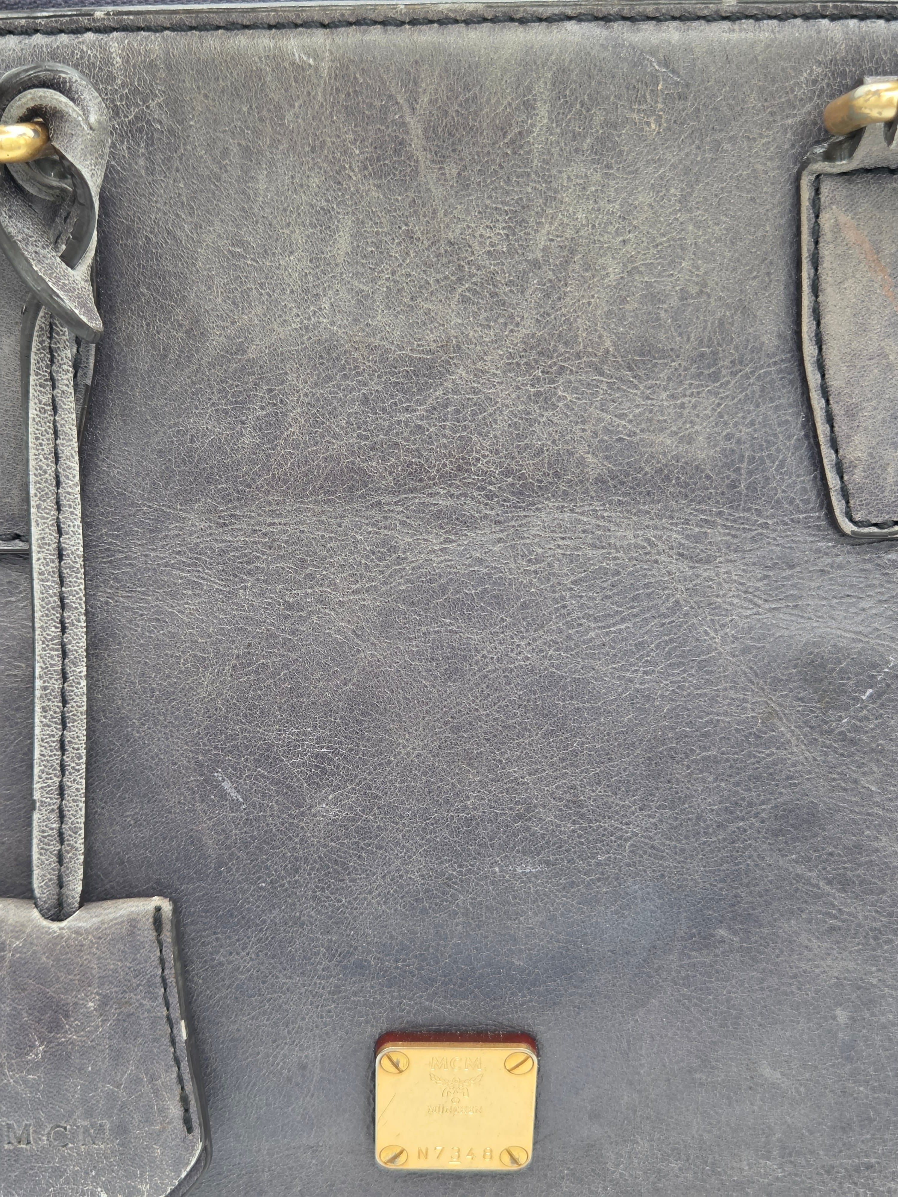 Close-up of a dark gray distressed leather bag showing the textured grain, gold-tone hardware, and a small gold-tone metal logo plaque at the bottom.