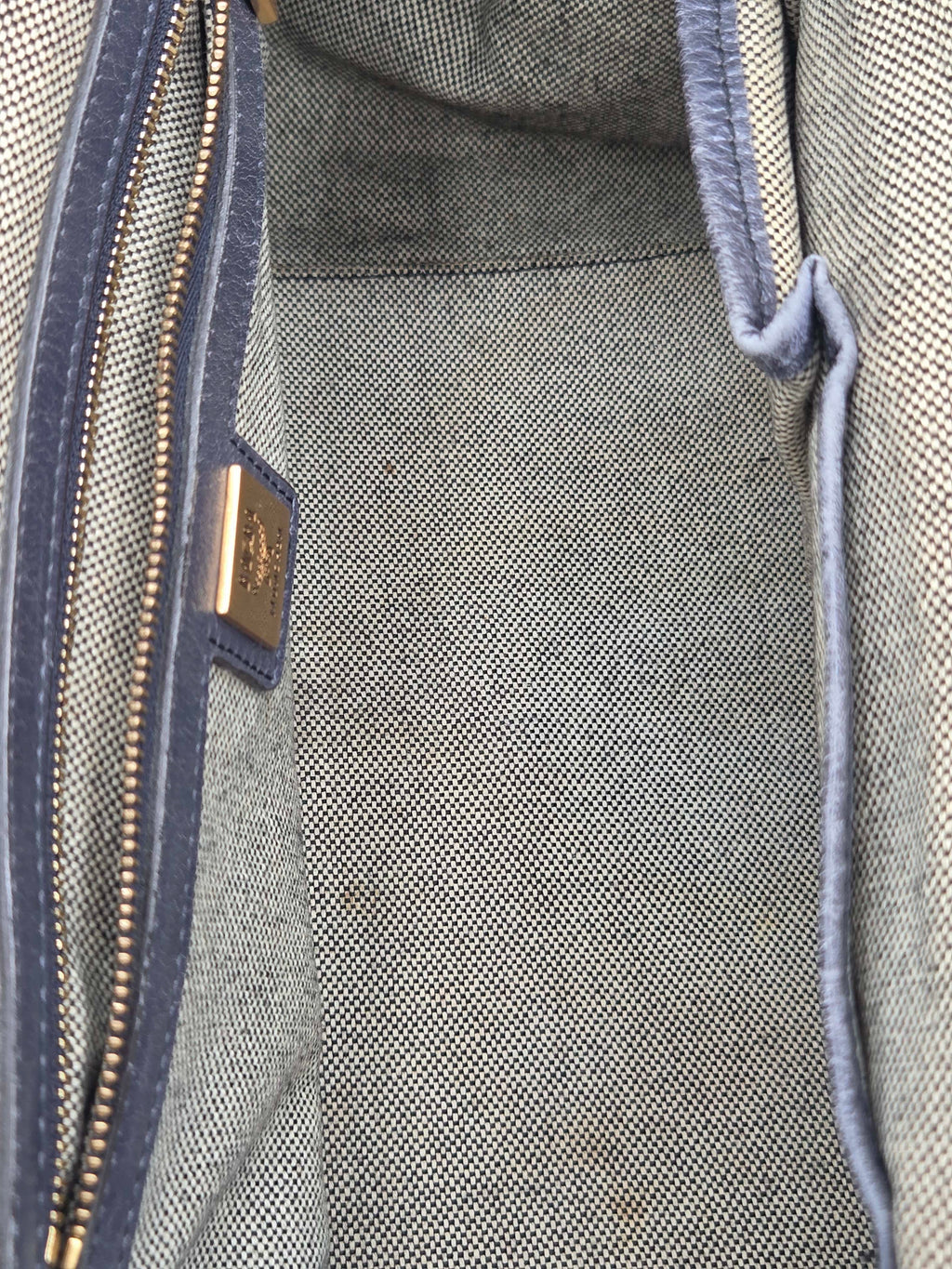 Close-up of the interior of a textured gray woven bag, showing a blue-gray zipper with a gold-tone metal pull tab and a small gold brand plaque.