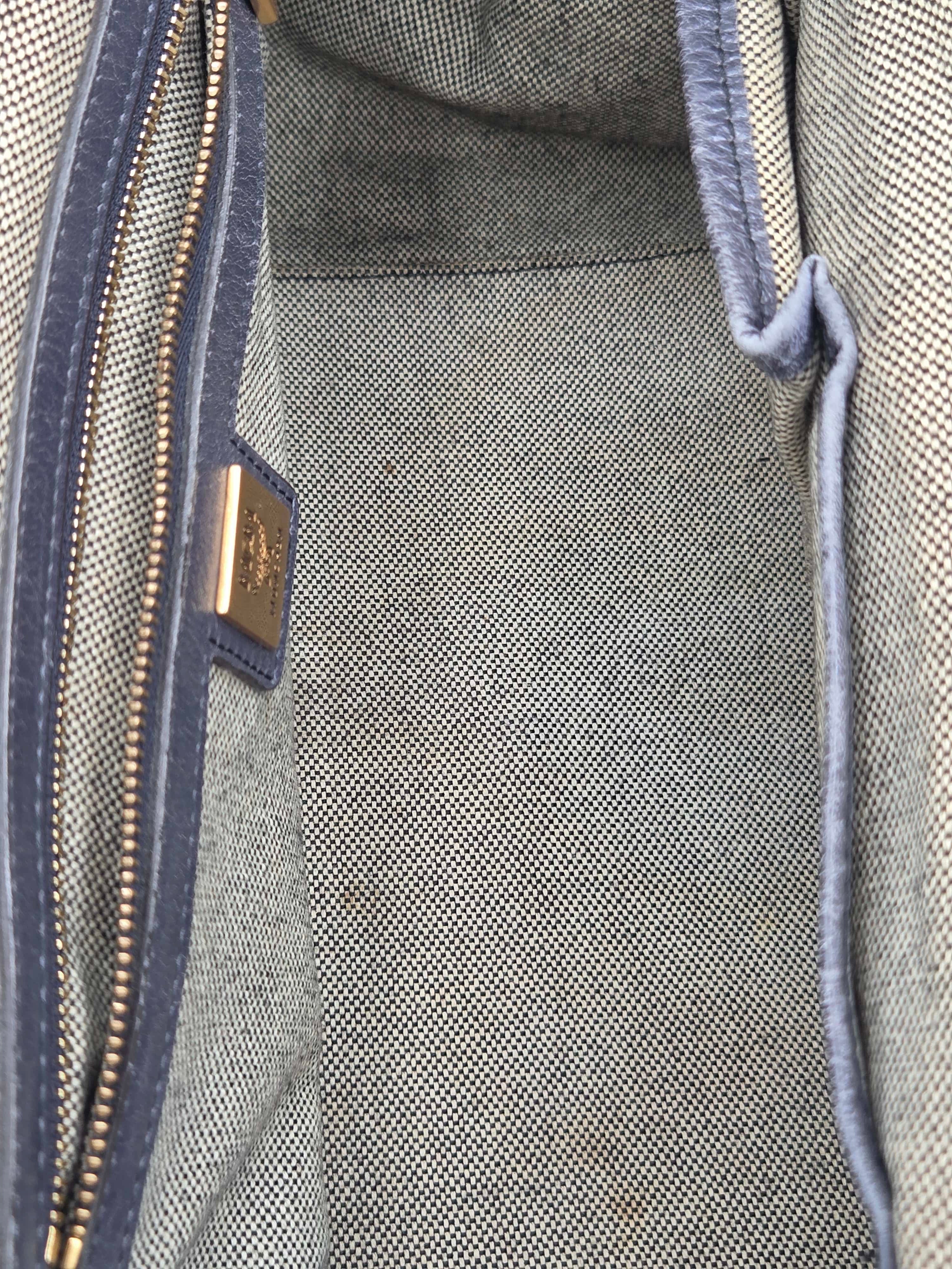 Close-up of the interior of a textured gray woven bag, showing a blue-gray zipper with a gold-tone metal pull tab and a small gold brand plaque.