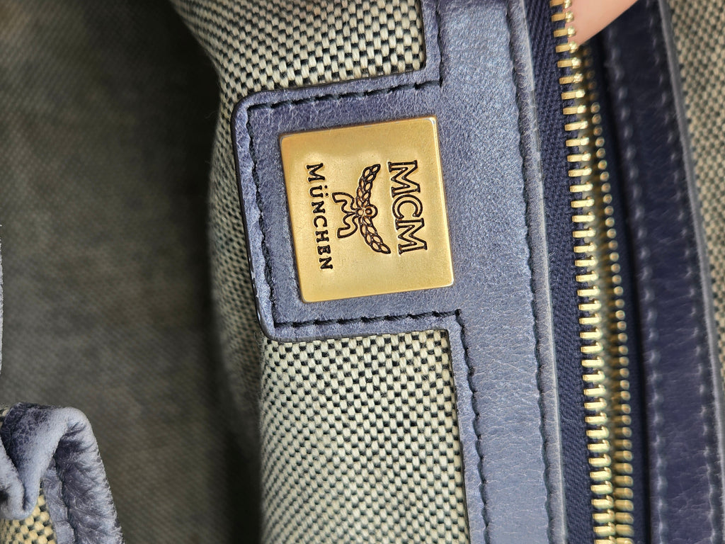 Close-up of an MCM München zipper pull tab with a gold-tone logo plate, set against a textured canvas and navy blue leather trim.
