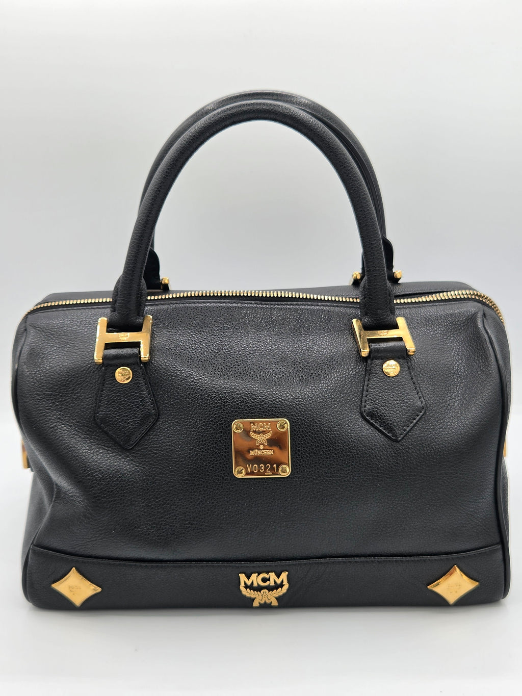 A black leather MCM handbag with gold-tone hardware, featuring dual top handles, a front zip closure, and the signature MCM logo plaque and monogram.
