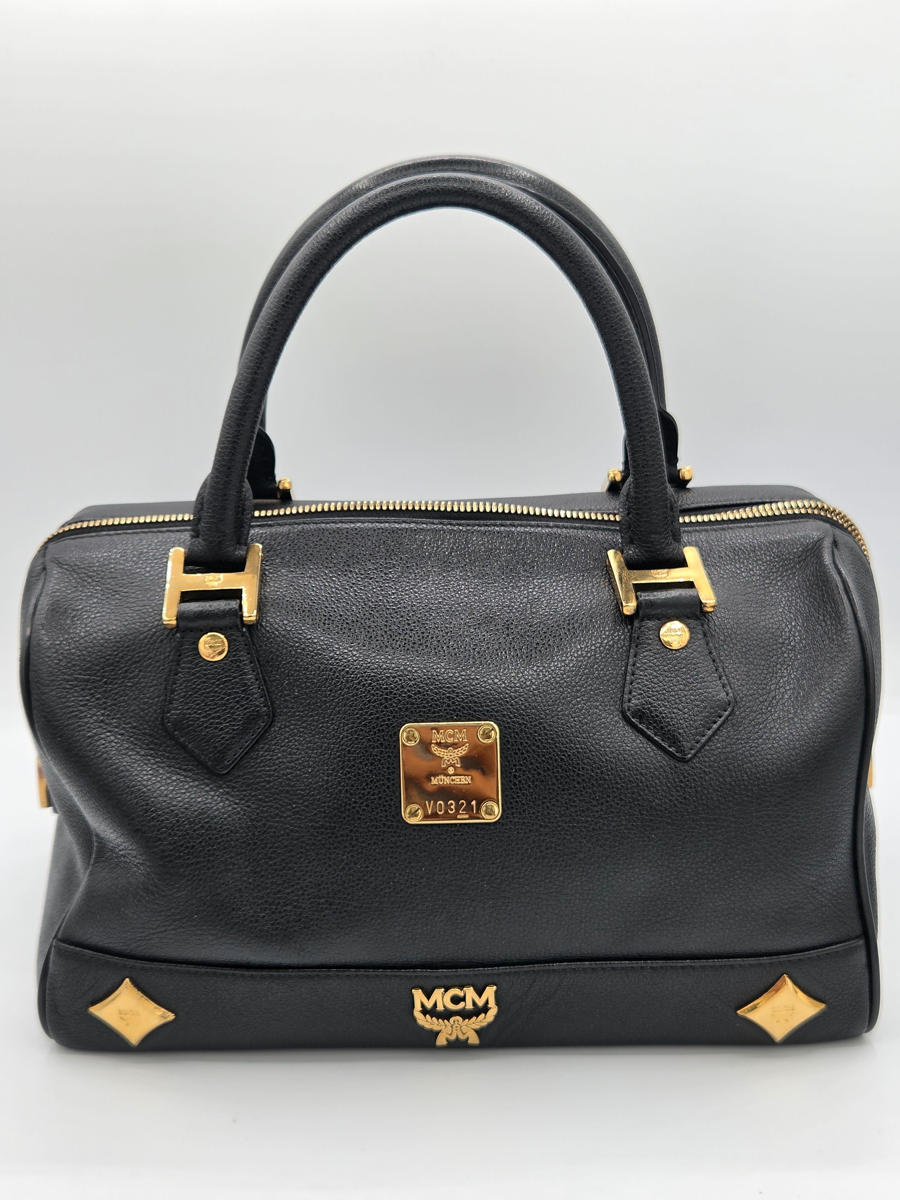 A black leather MCM handbag with gold-tone hardware, featuring dual top handles, a front zip closure, and the signature MCM logo plaque and monogram.