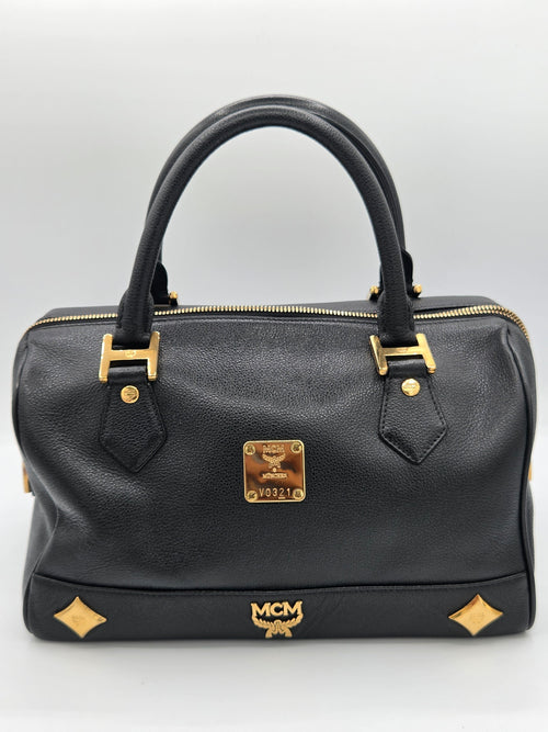 A black leather MCM handbag with gold-tone hardware, featuring dual top handles, a front zip closure, and the signature MCM logo plaque and monogram.