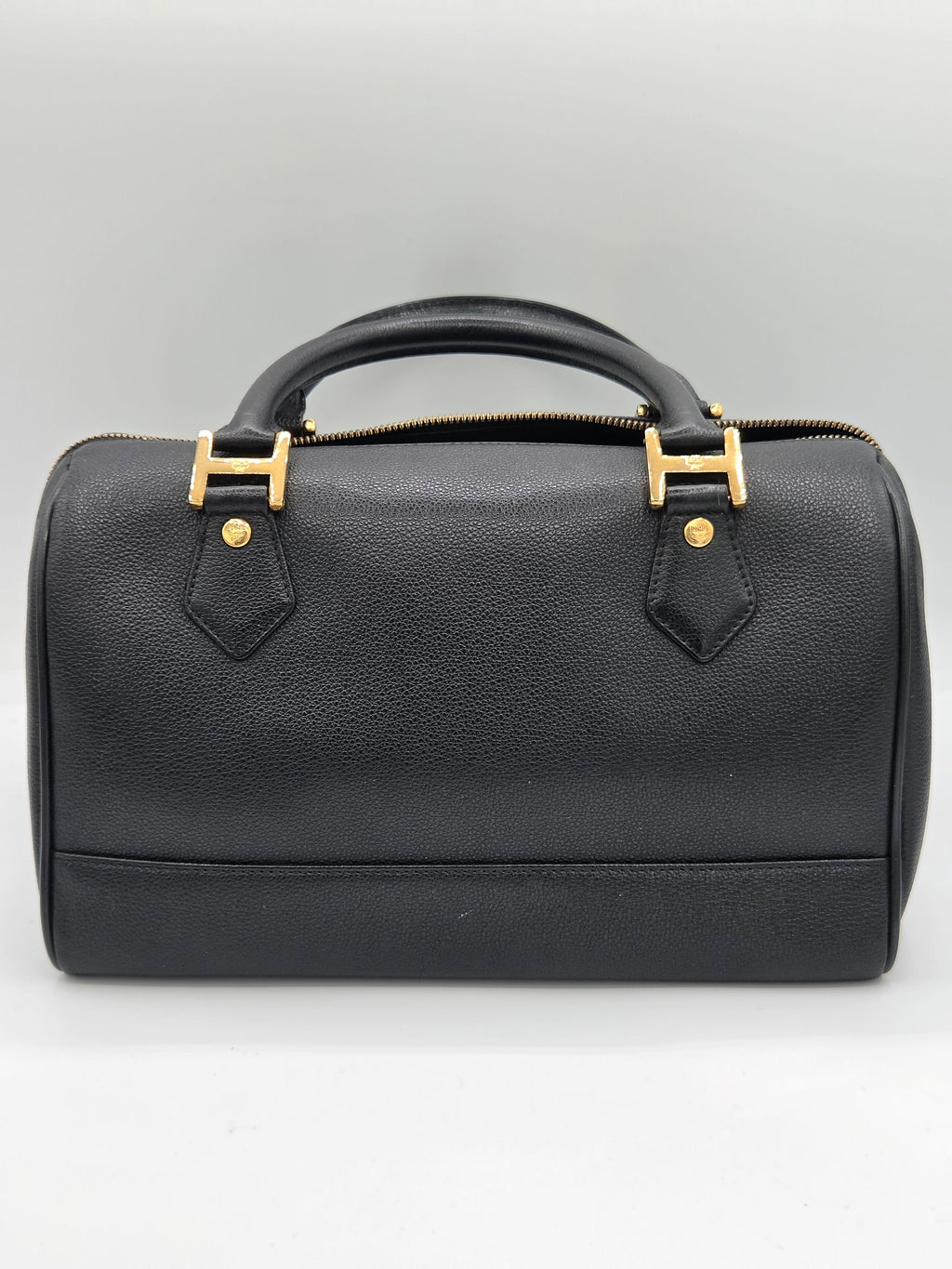 A black pebbled leather handbag with gold-tone hardware, featuring a top zipper closure and a curved top handle, shown from a top-down angle on a light gray background.