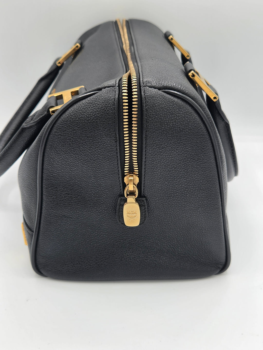 A black pebbled leather handbag shown from an angled top-down view, featuring a prominent gold zipper with a gold-tone pull tab and gold hardware details.