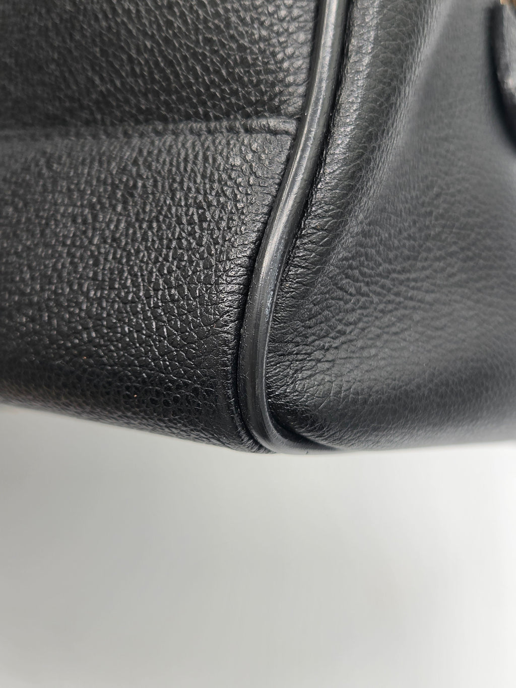 Close-up detail of a black pebbled leather bag corner showing the grain texture and a thin dark edge trim against a light gray background.