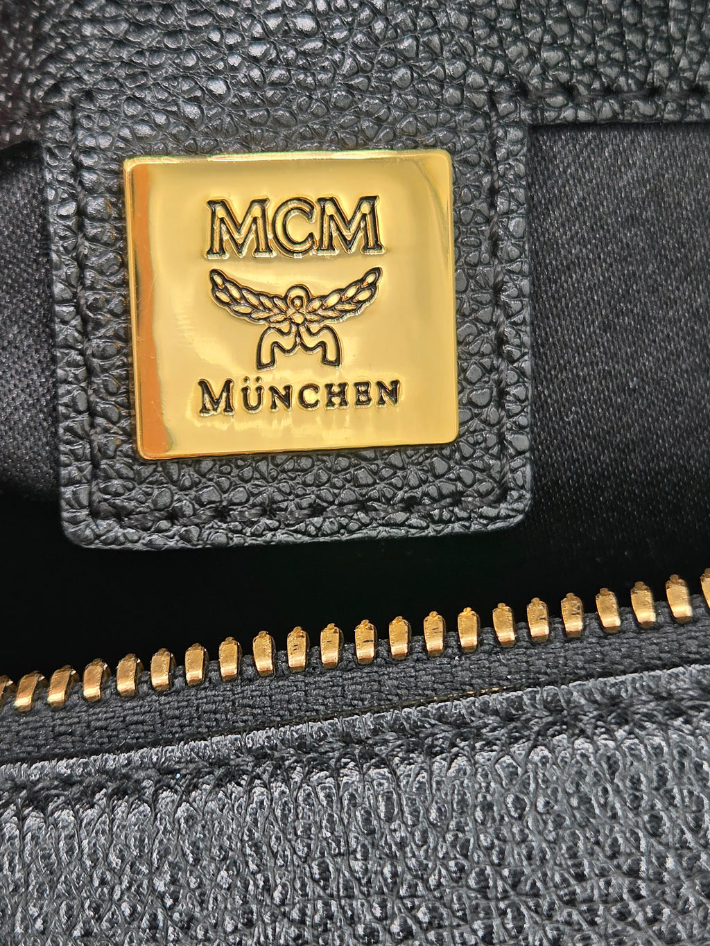 Close-up of an MCM München brand patch on a black textured leather surface next to a gold zipper.