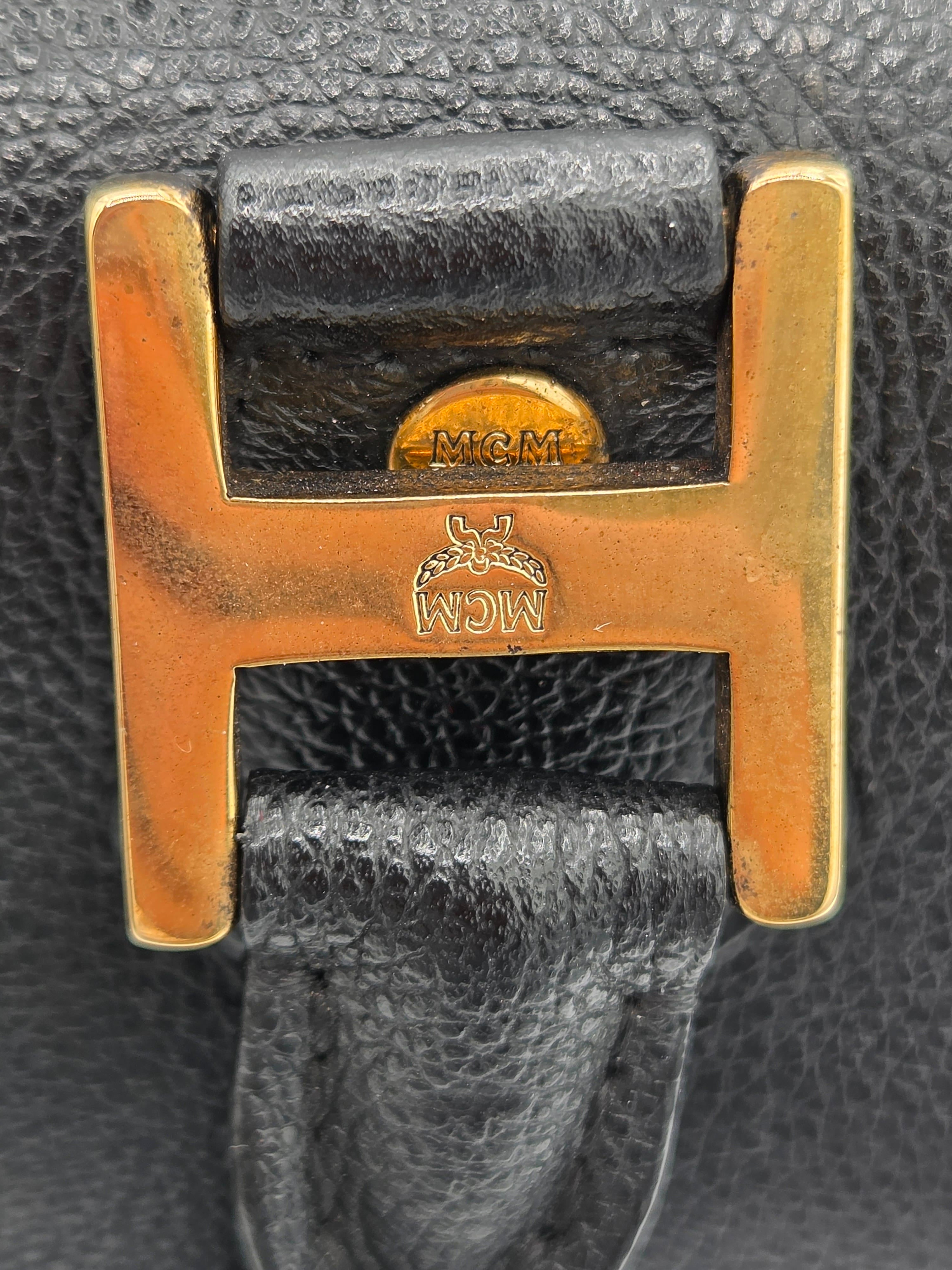 Close-up of an MCM black leather strap with a polished gold-tone H-buckle and brand stamp.