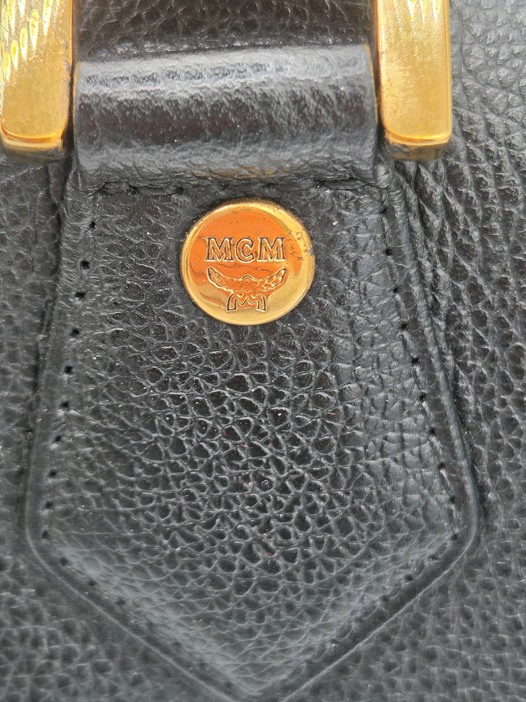 Close-up of a black pebbled leather accessory with a round gold-tone metal emblem engraved with the MCM logo, alongside gold-tone hardware.