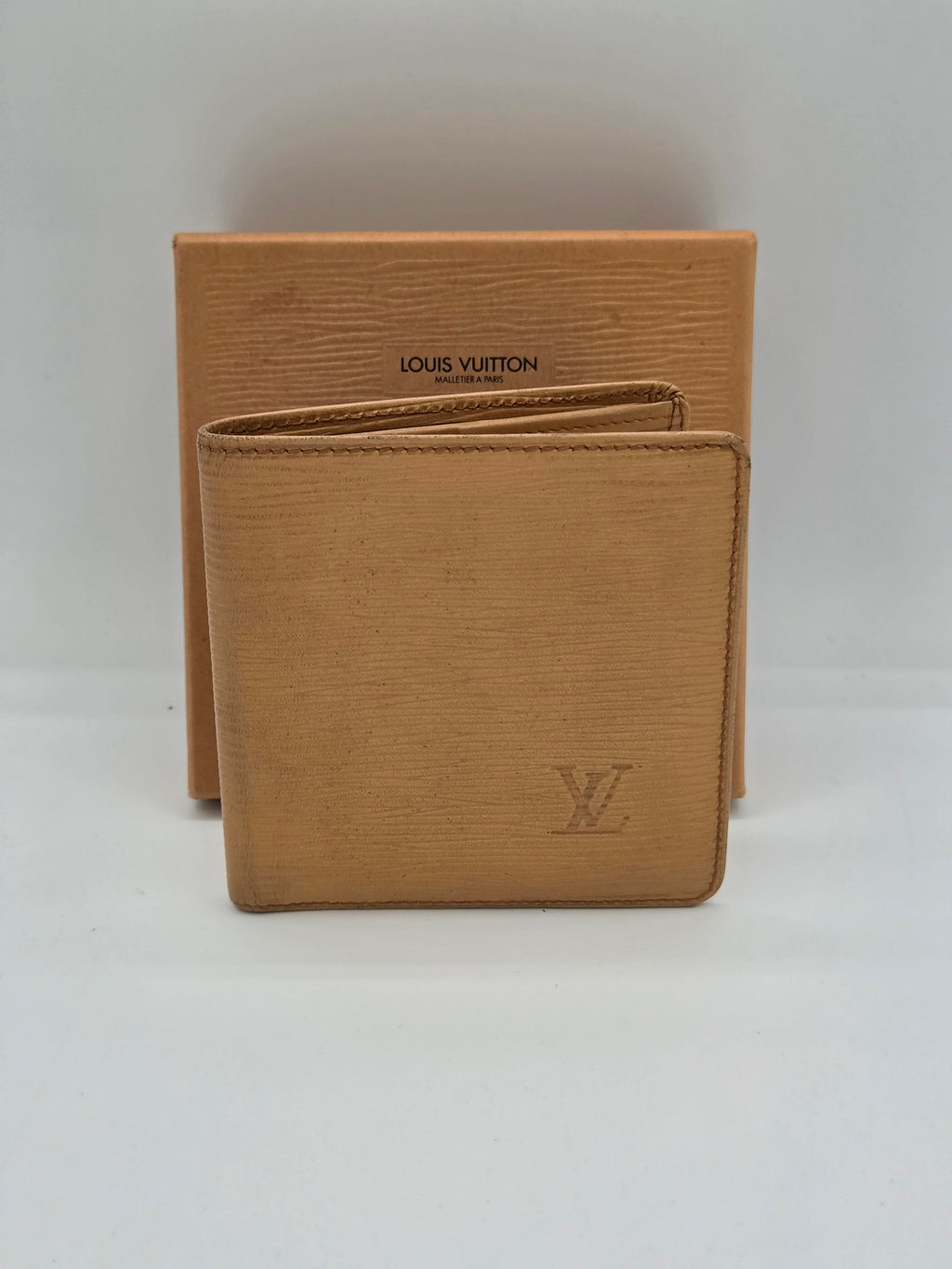 A Louis Vuitton tan leather bifold wallet with an embossed LV logo, resting on top of its matching tan textured box against a plain white background.