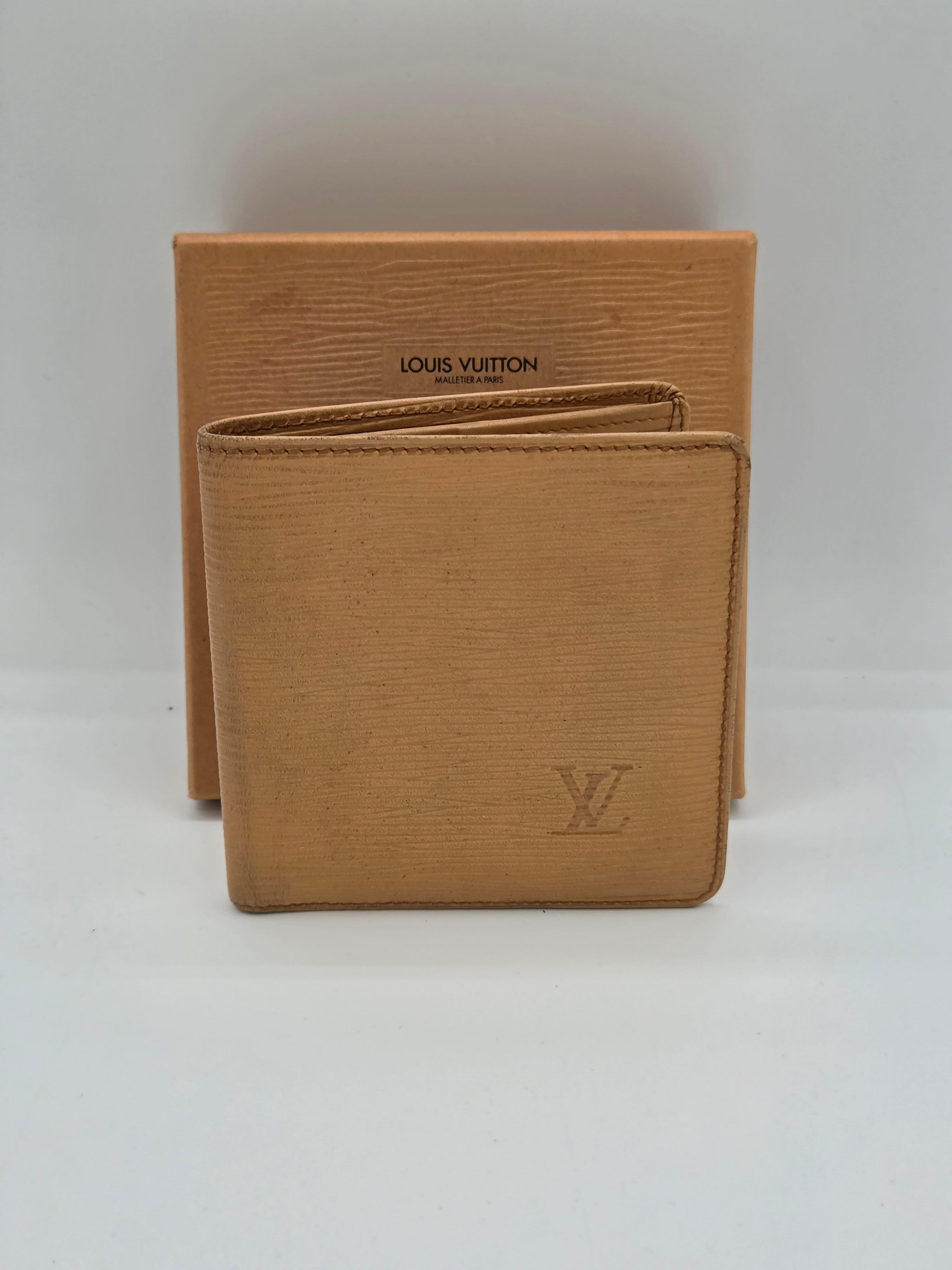 A Louis Vuitton tan leather bifold wallet with an embossed LV logo, resting on top of its matching tan textured box against a plain white background.