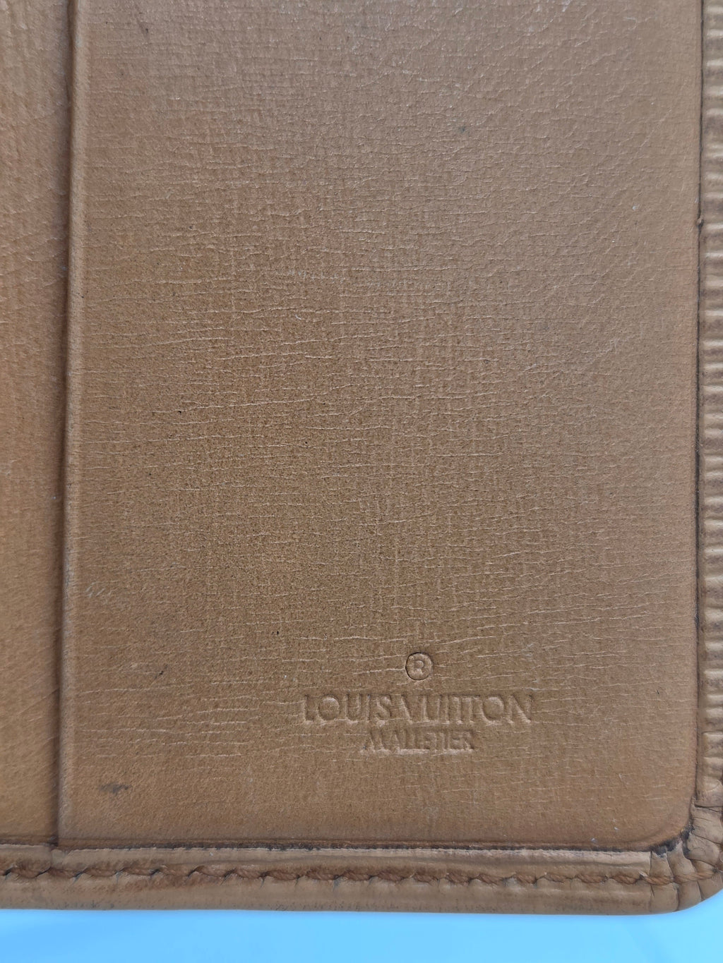 Close-up of a tan textured leather item, likely a wallet or document holder, with a debossed Louis Vuitton monogram on the right side.