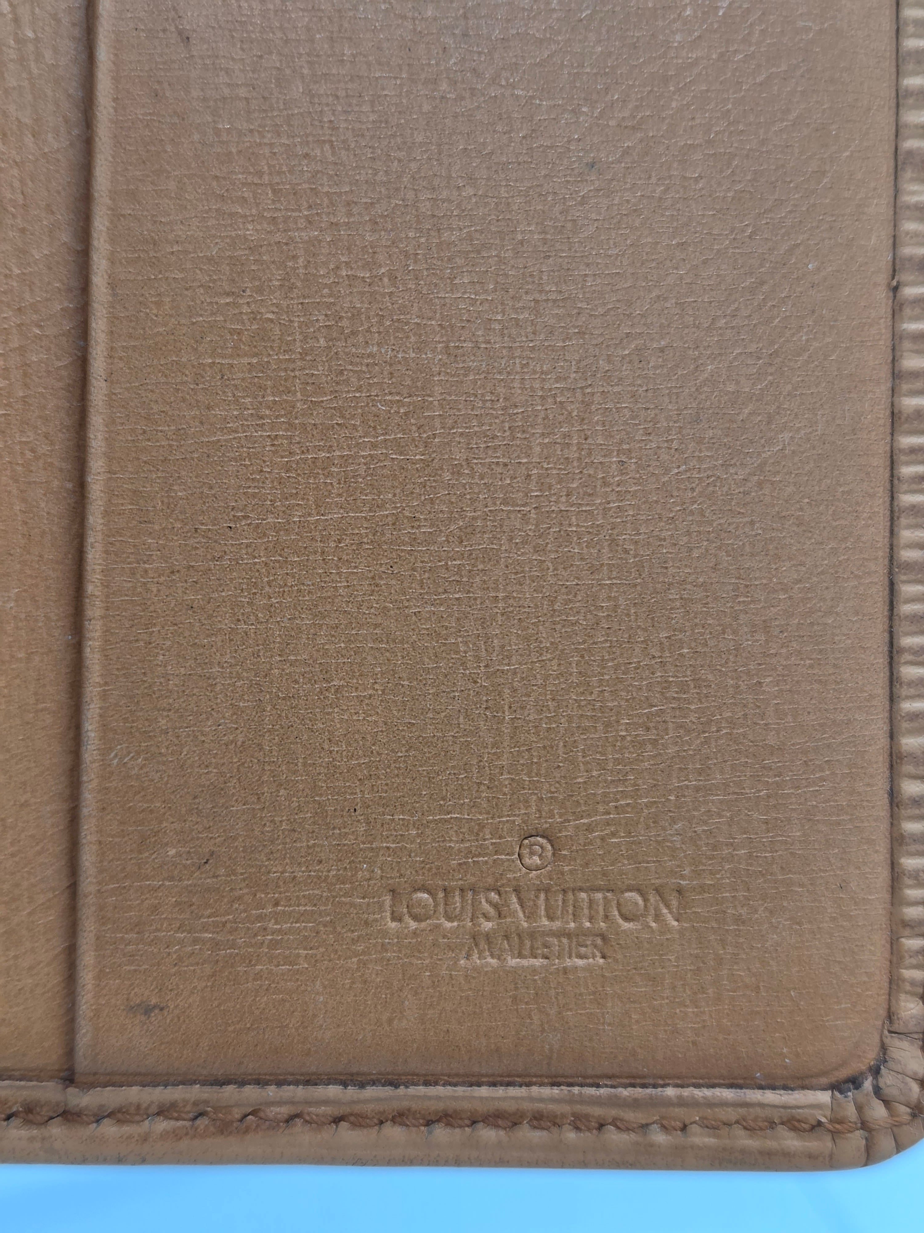 Close-up of a tan textured leather item, likely a wallet or document holder, with a debossed Louis Vuitton monogram on the right side.