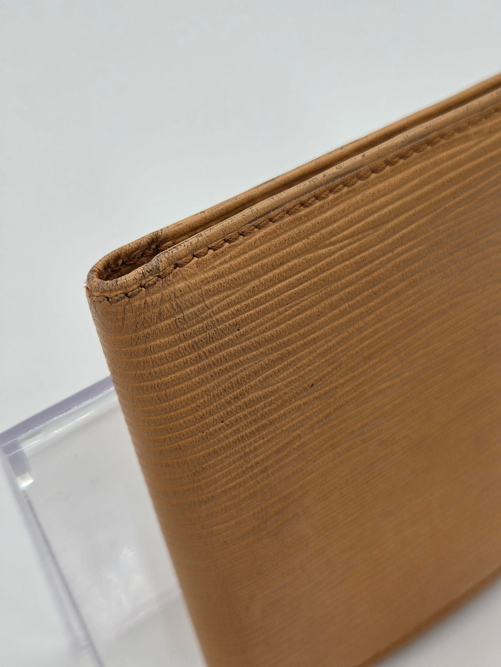 A close-up of a tan textured leather item, showing fine grain and stitched edges, resting on a clear acrylic stand against a white background.