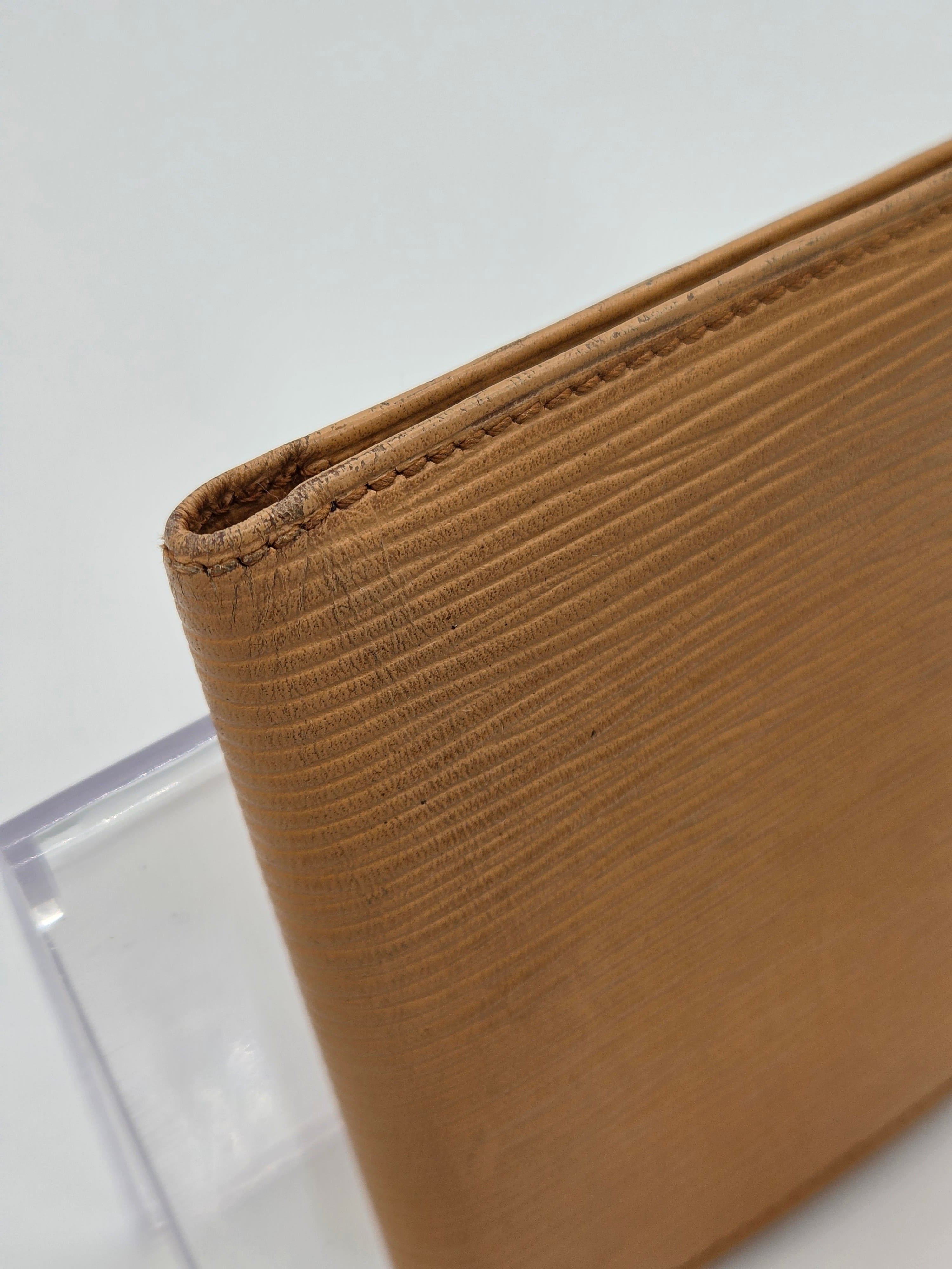 A close-up of a tan textured leather item, showing fine grain and stitched edges, resting on a clear acrylic stand against a white background.