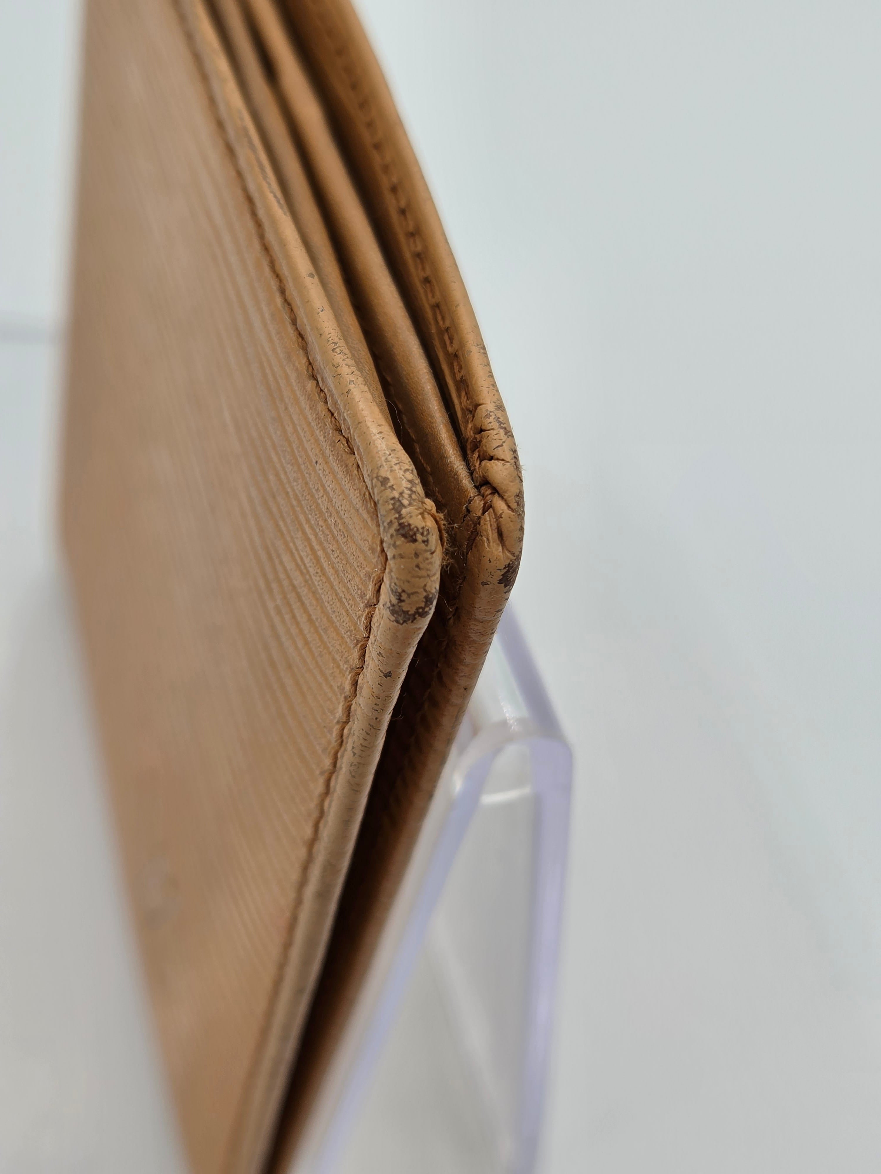Close-up of a tan leather wallet corner showing the stitched edge and a clear protective plastic cover on a white background.