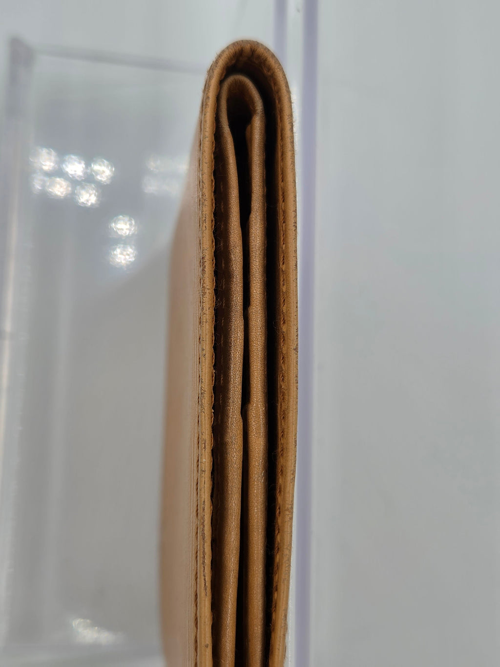 A close-up side view of a tan leather wallet or card holder, showing the layered construction and edge stitching, resting on a clear acrylic stand against a white background.