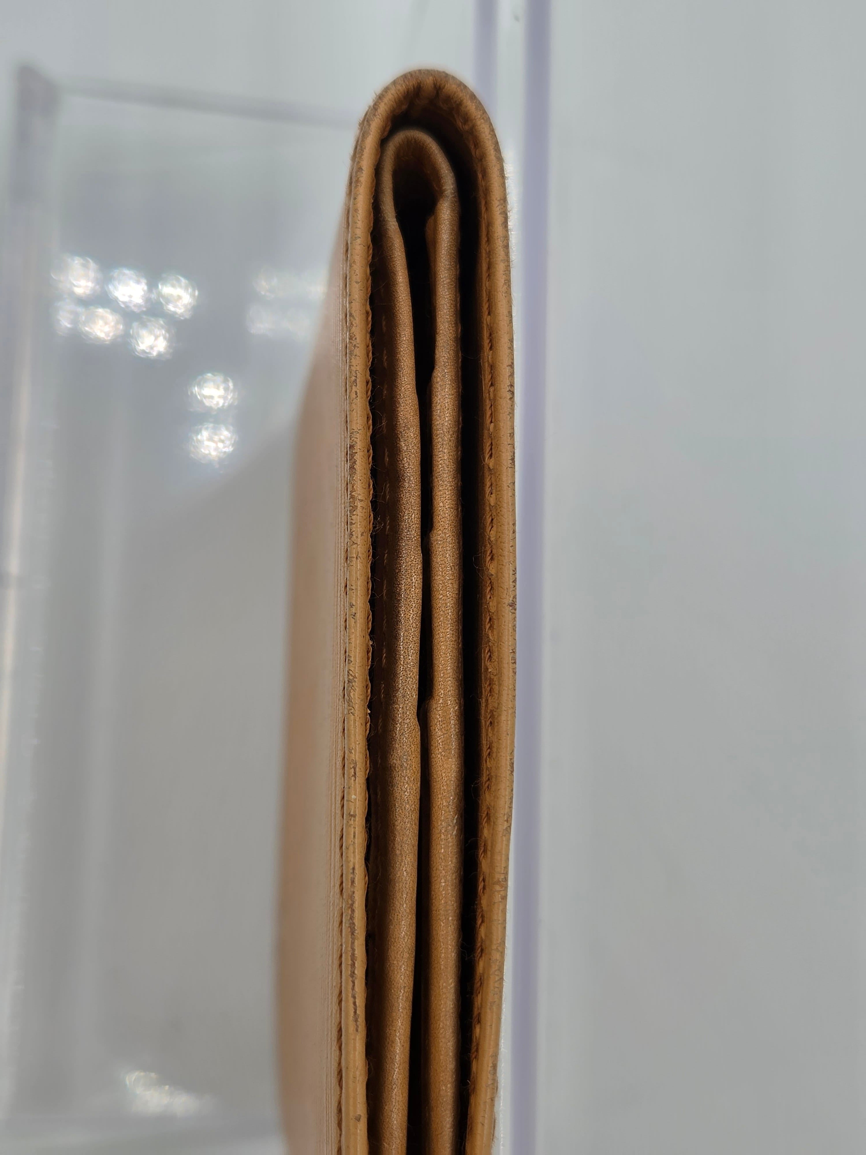 A close-up side view of a tan leather wallet or card holder, showing the layered construction and edge stitching, resting on a clear acrylic stand against a white background.