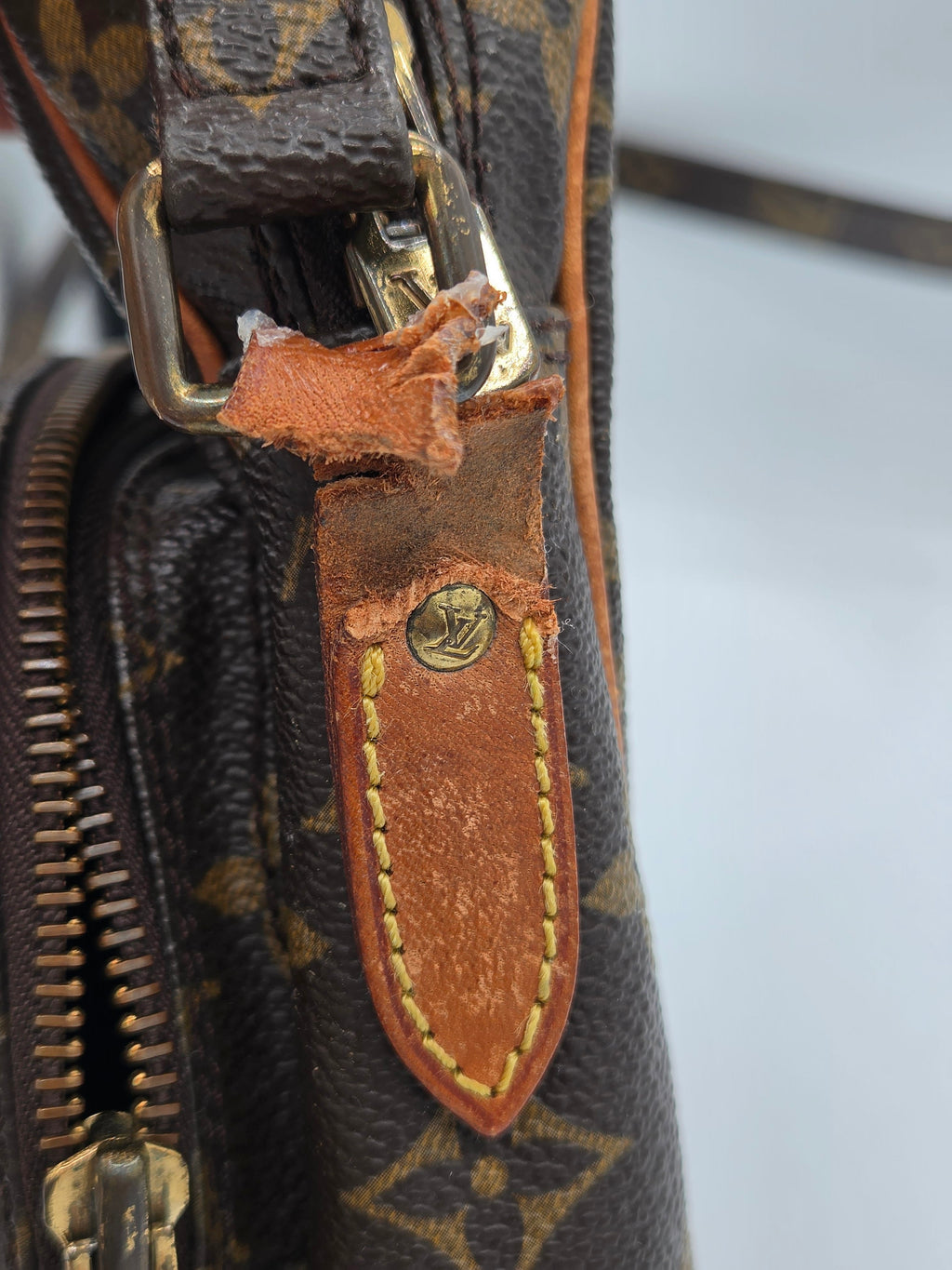 Close-up of a Louis Vuitton monogram bag showing a worn brown leather tab with a damaged torn edge and a brass screw, alongside gold-tone hardware and zipper.