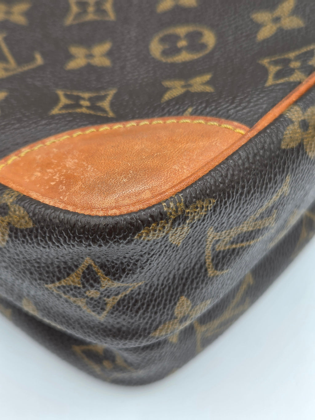 Close-up of a Louis Vuitton monogram canvas bag corner showing the dark brown coated canvas with tan monogram motifs and a tan leather trim piece.