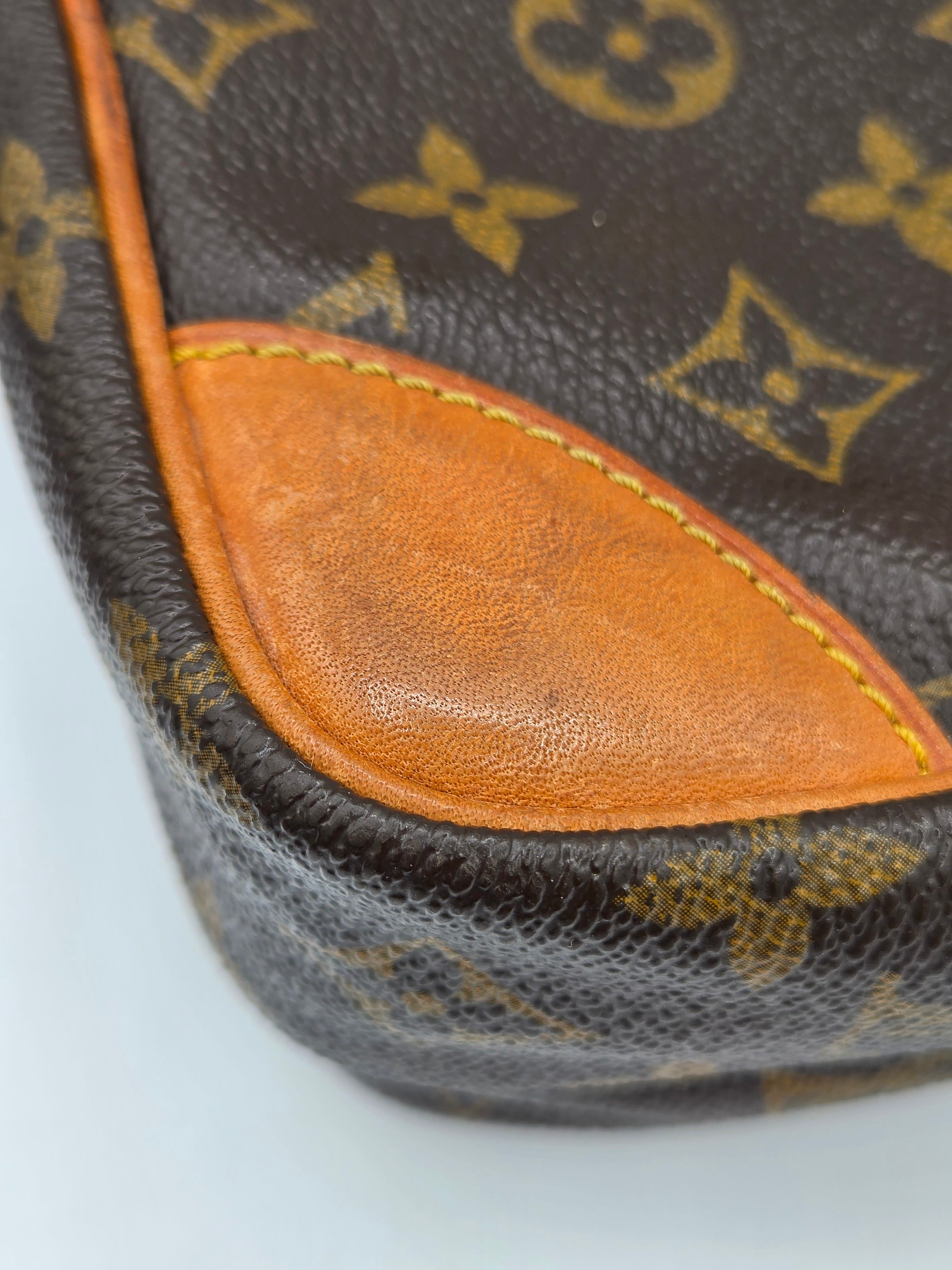 Close-up of a Louis Vuitton monogram canvas bag corner showing the dark brown coated canvas with gold monogram print and a contrasting orange-brown leather trim.