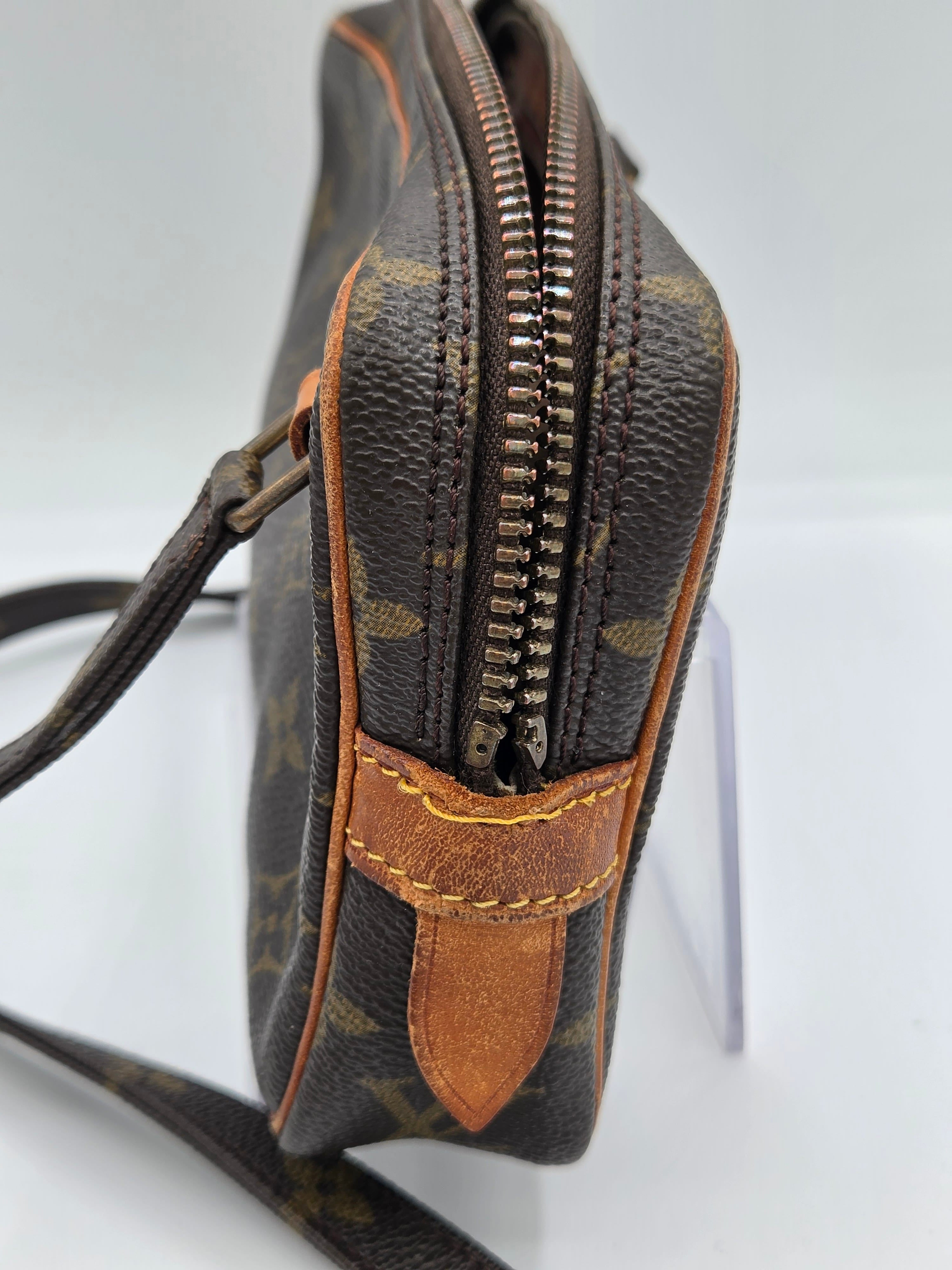 A close-up of a Louis Vuitton monogram crossbody bag showing the dark brown coated canvas with its signature tan and green logo print, a brown leather tab, and a silver-toned zipper.