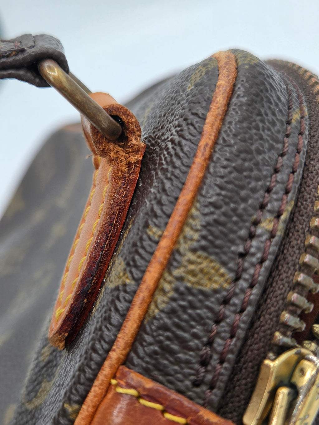 Close-up of a dark brown monogram handbag showing the textured coated canvas, tan leather trim, brass zipper pull, and a worn metal loop attachment.