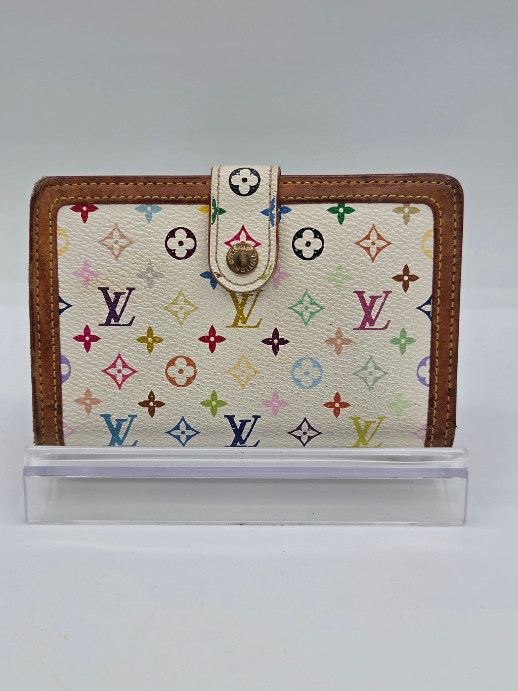 A Louis Vuitton monogram canvas passport cover or wallet with brown leather trim and a snap strap closure, shown standing upright next to a clear plastic protective sleeve on a light gray background.