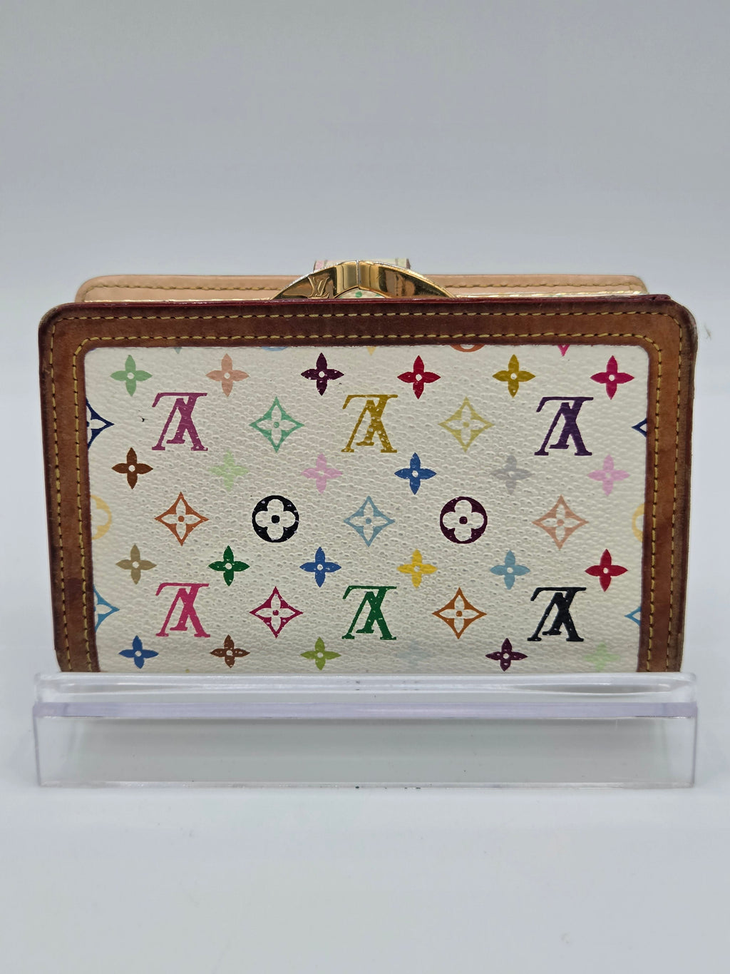 Louis Vuitton monogram canvas clutch wallet with brown leather trim and a gold-tone clasp, shown next to a clear plastic protective sleeve on a light gray background.