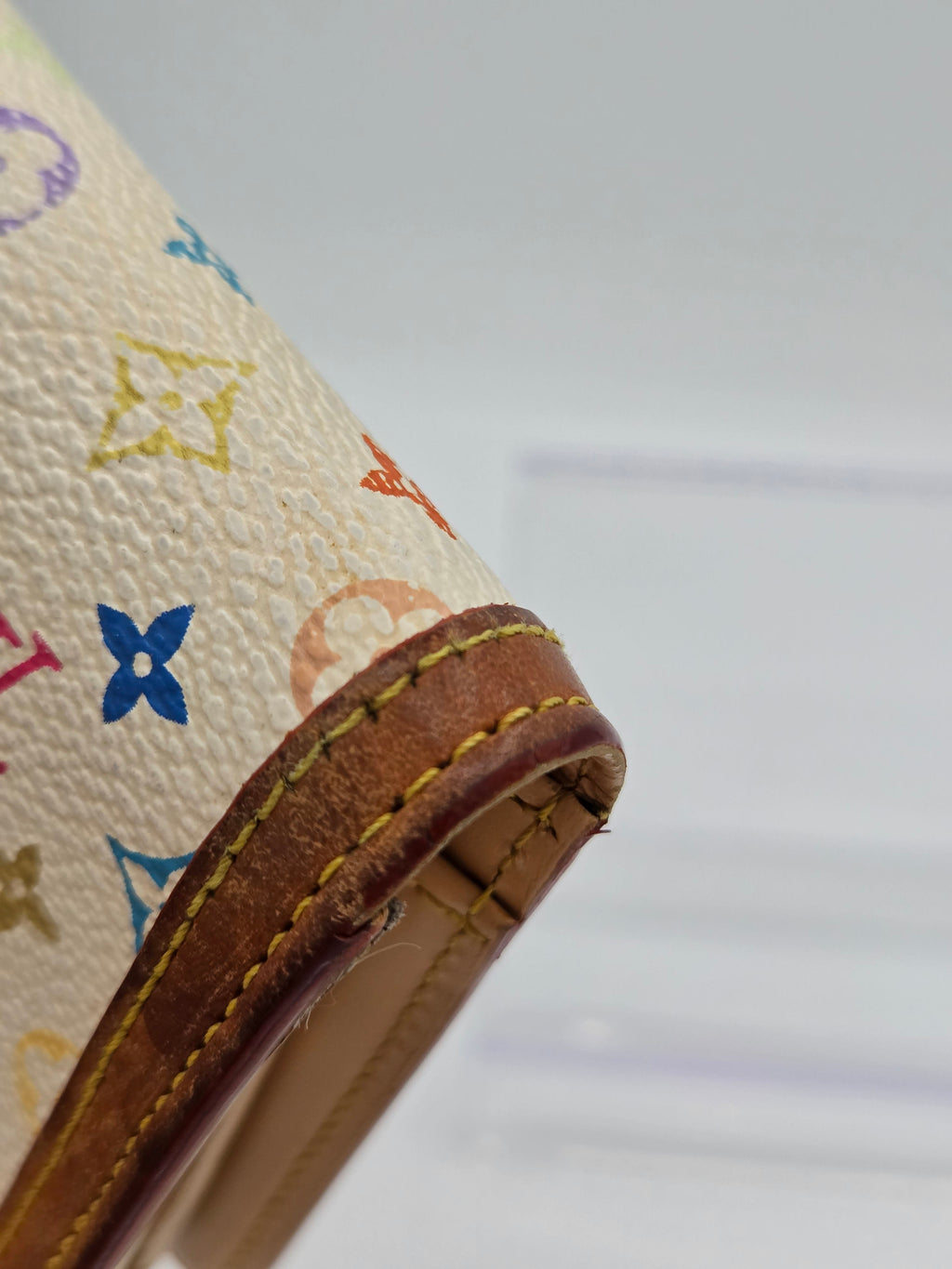 Close-up of a Louis Vuitton monogram canvas handbag corner with a brown leather trim and yellow stitching, showing the cream canvas base with multicolor monogram motifs.