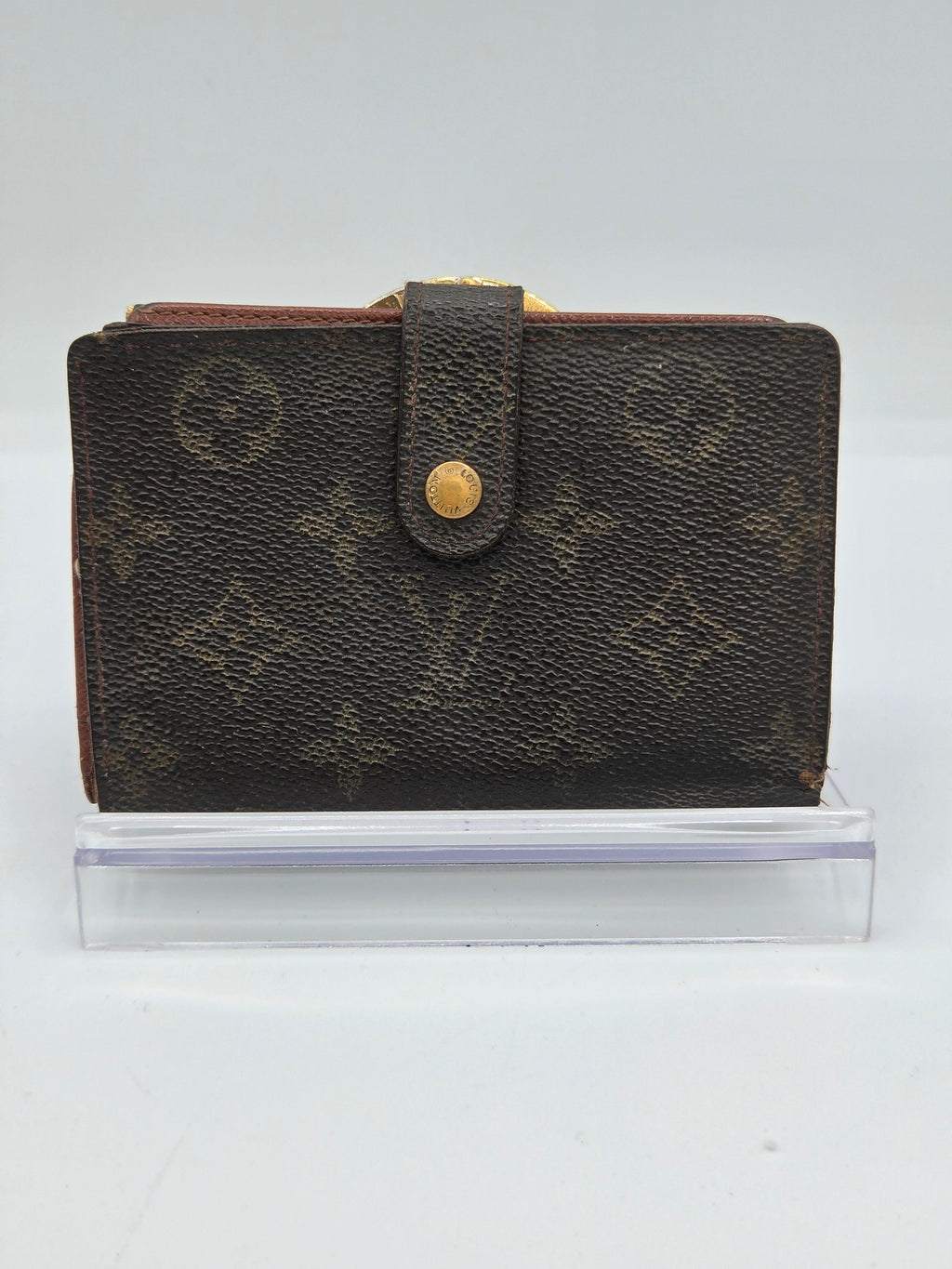 A Louis Vuitton monogram notebook or planner cover in dark brown coated canvas with gold-tone hardware, shown standing upright next to a clear acrylic block against a white background.